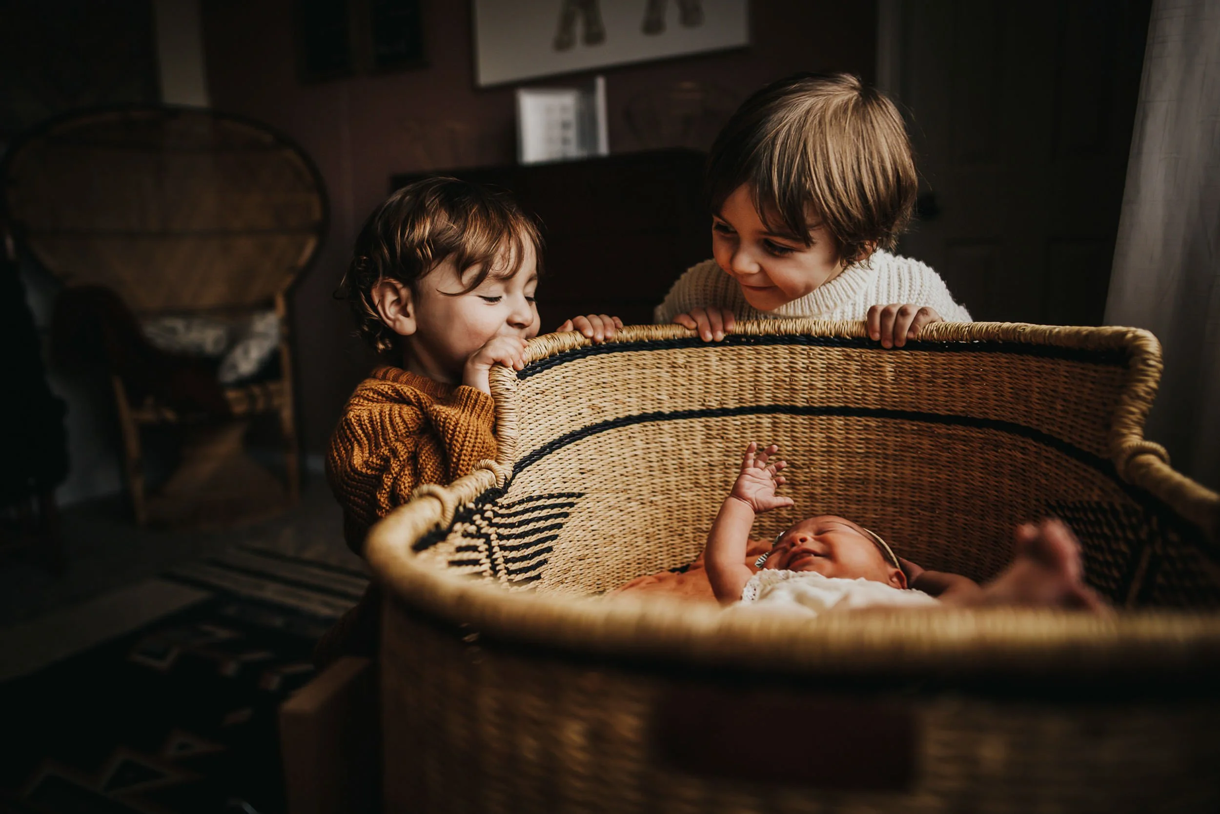 Two young children lean over and peek into a wicker basket together indoors.