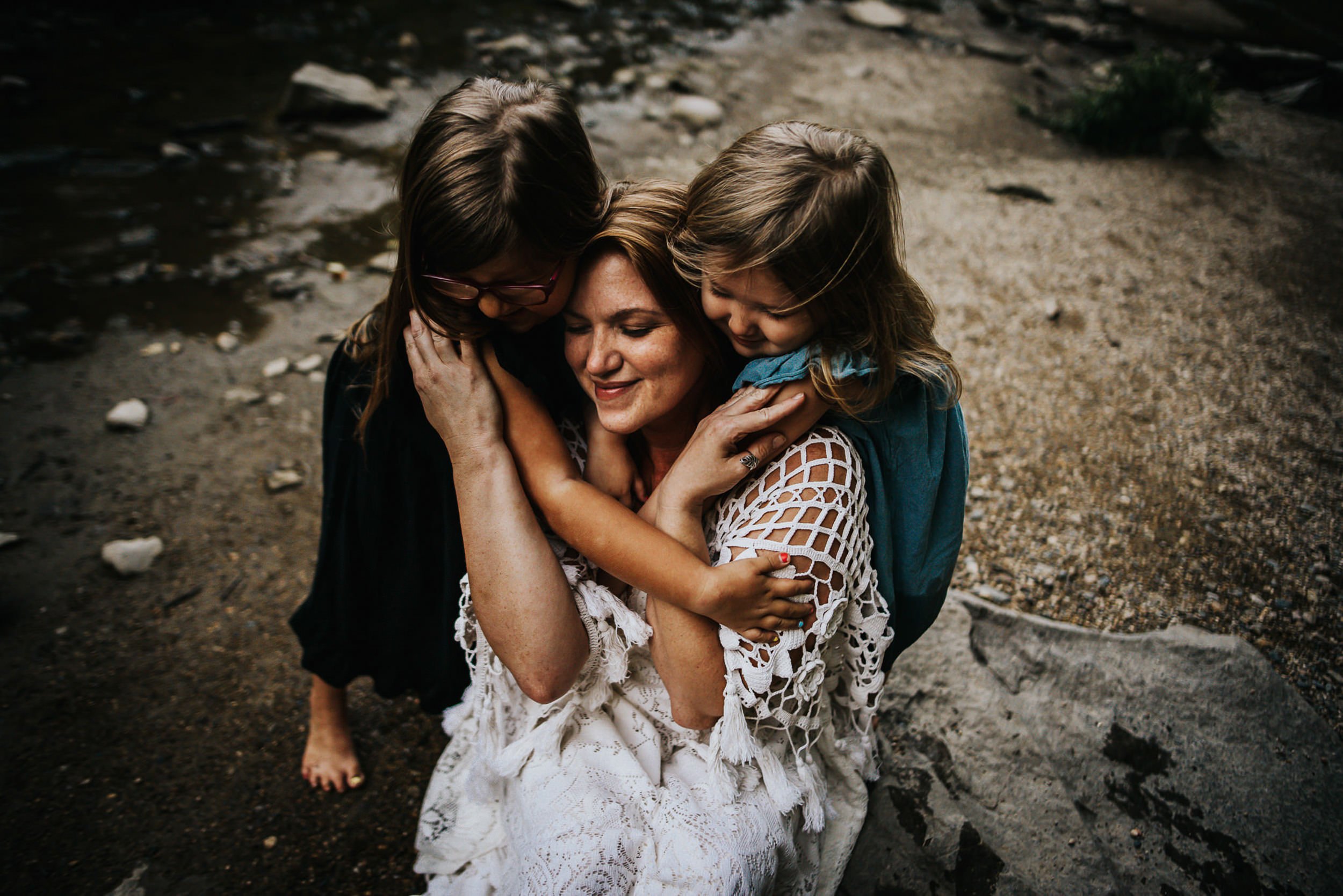 Mom in cream crochet top embracing two daughters, one in teal, by a rocky creek during a family session.