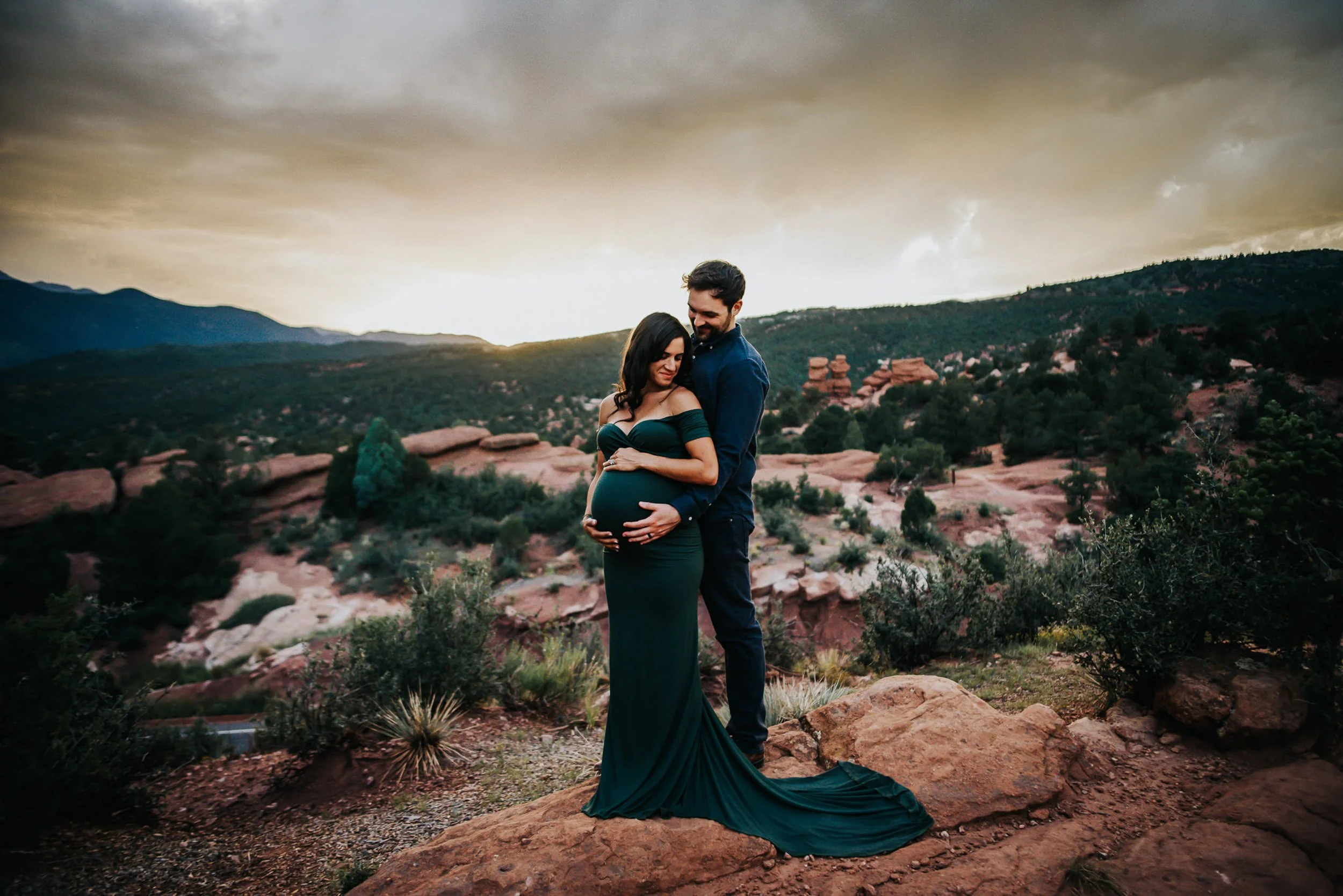 Couple in coordinated deep teal and charcoal standing together with dramatic Colorado mountain backdrop at golden hour.