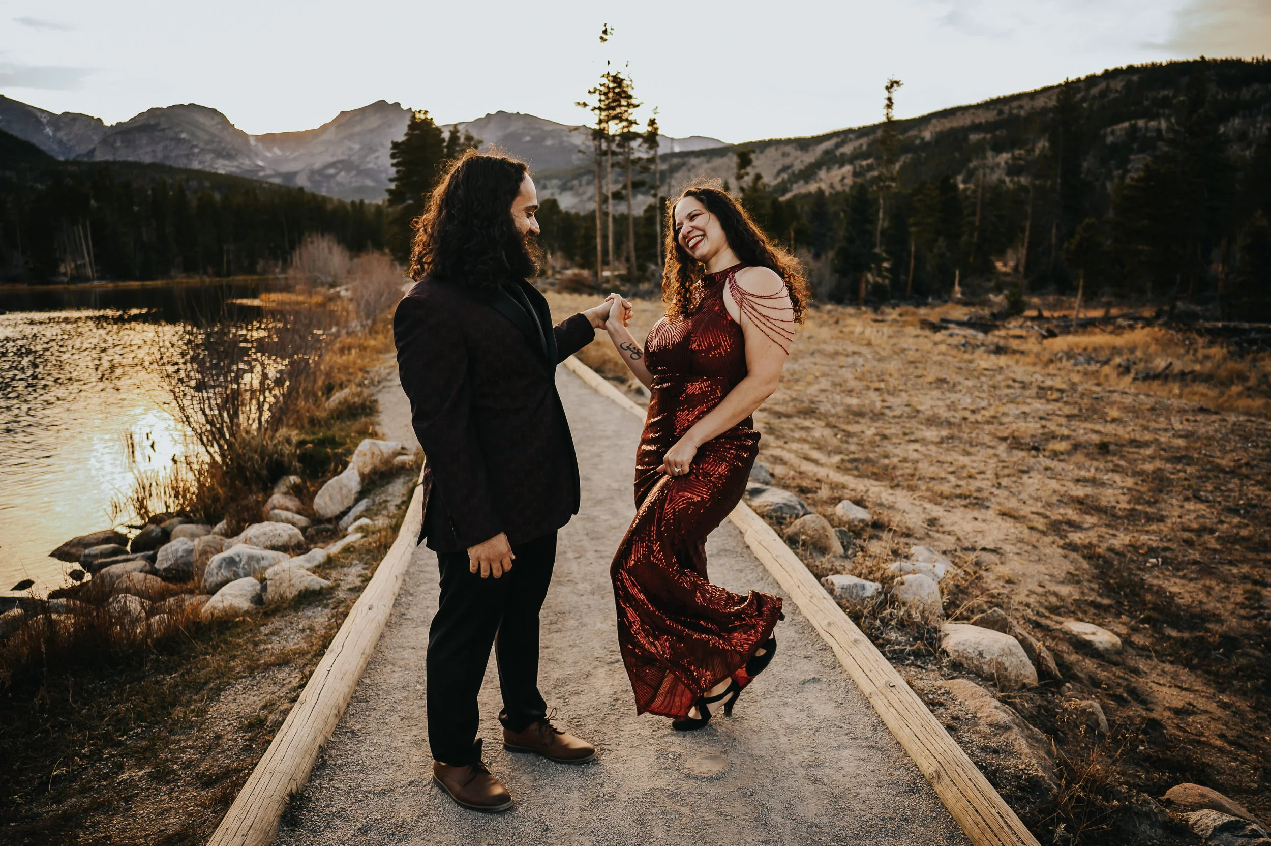 A bride in a red sequined gown and her groom dance and laugh together on a mountain trail.