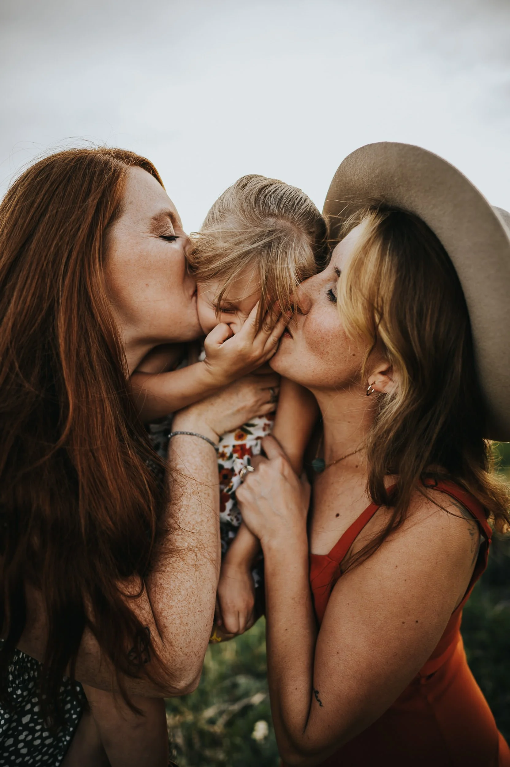 Two mothers hold their daughter between then as they sport complimentary colors and a wide brimmed hat for texture.