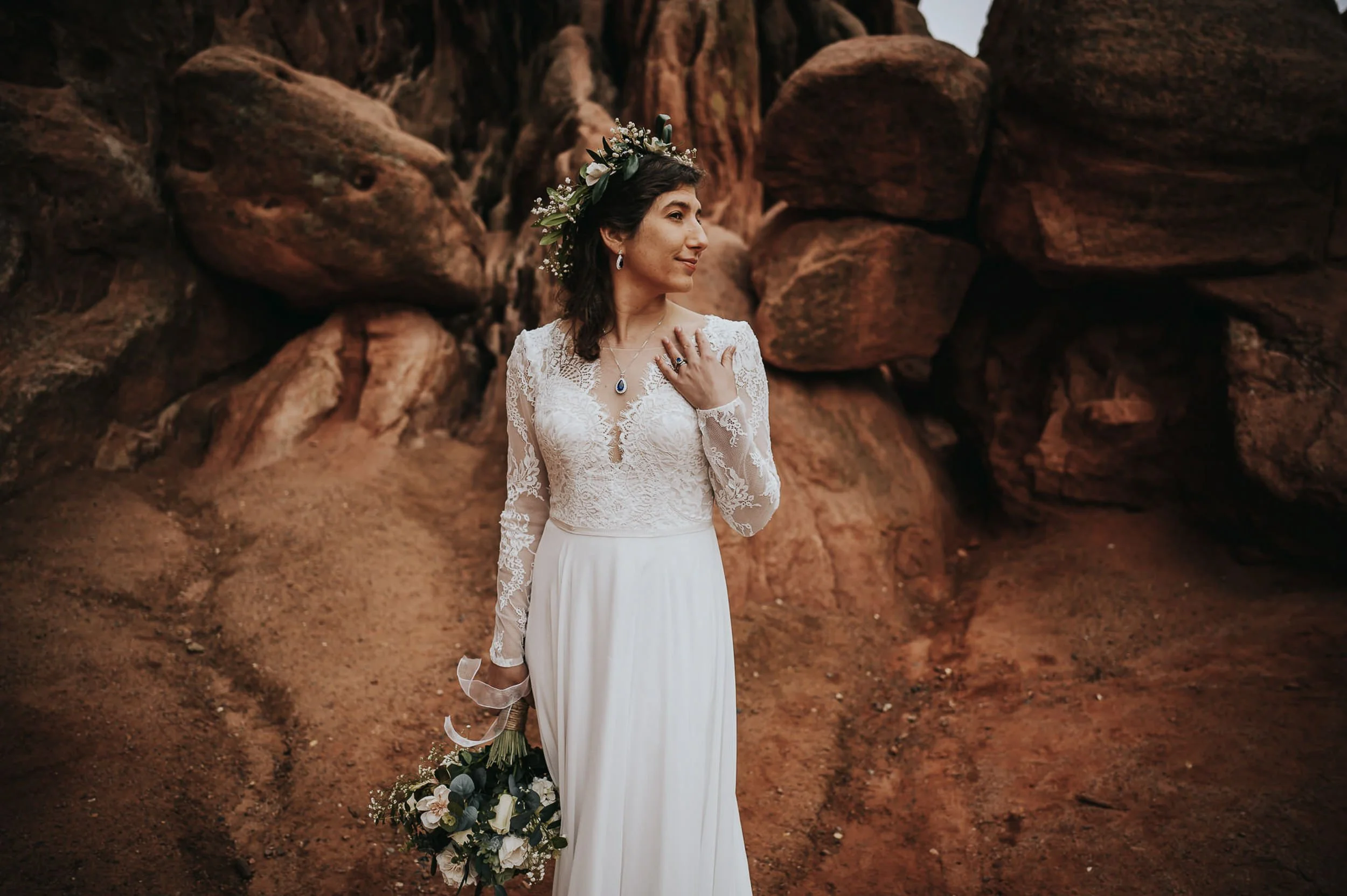 A bride in a lace gown and flower crown stands alone among red rocks, looking down softly, Colorado landscape around her.