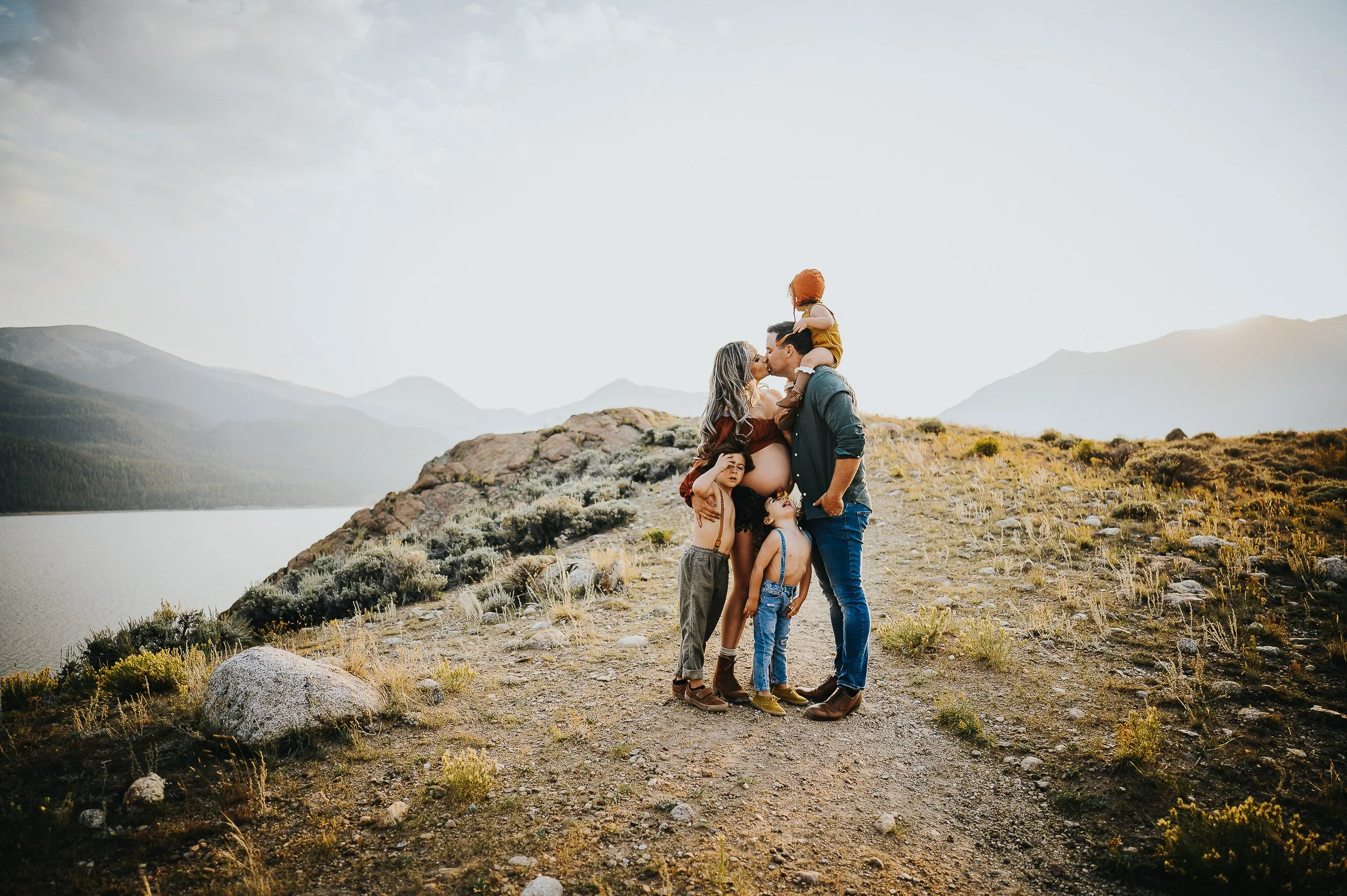Family of four at a Colorado mountain lake, parents sharing a kiss, toddler on dad's shoulders in mustard, coordinated in tan and navy.