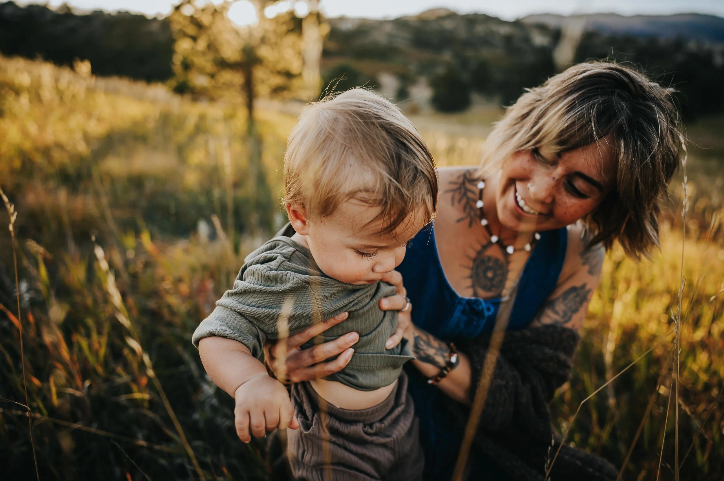 A toddler boy reaches toward his smiling mom in a golden autumn field in Colorado.