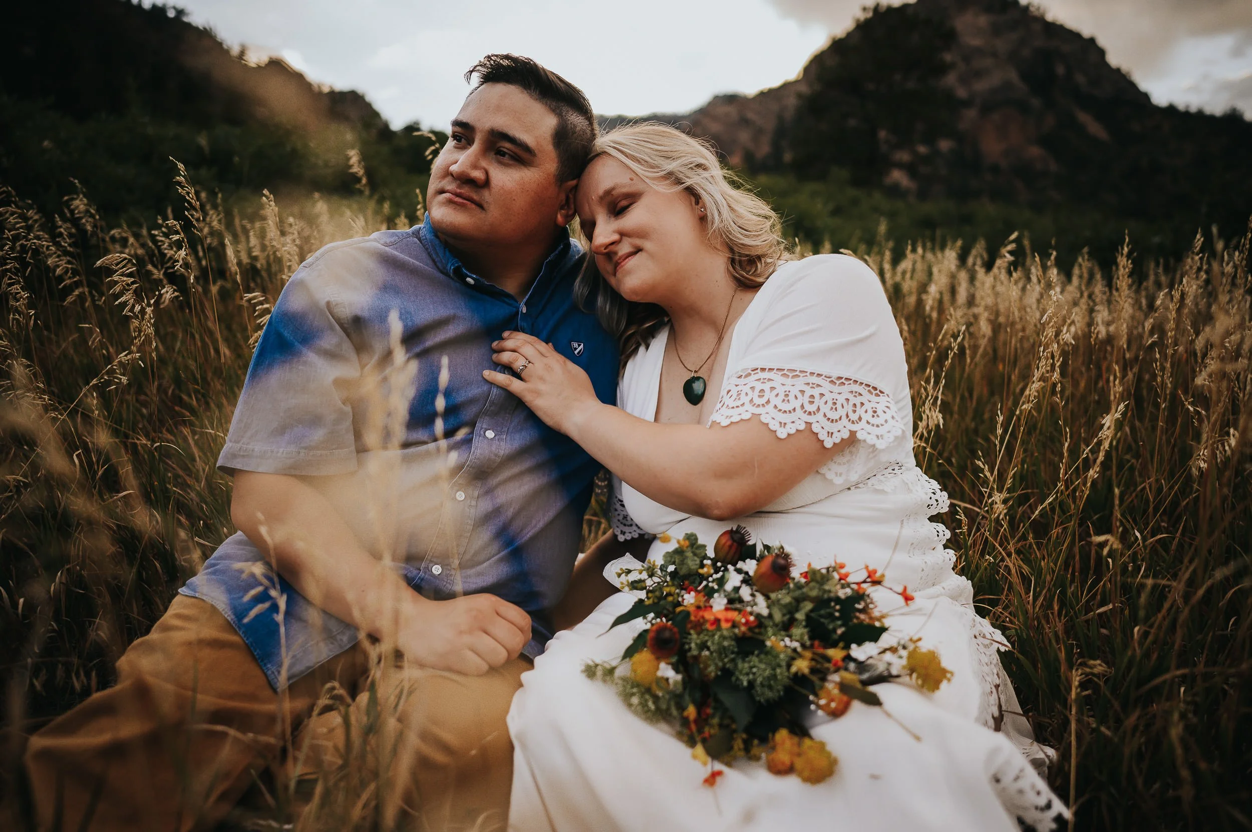 A couple sits together in a Colorado mountain field, she in a white lace dress with a bright wildflower bouquet in her lap.