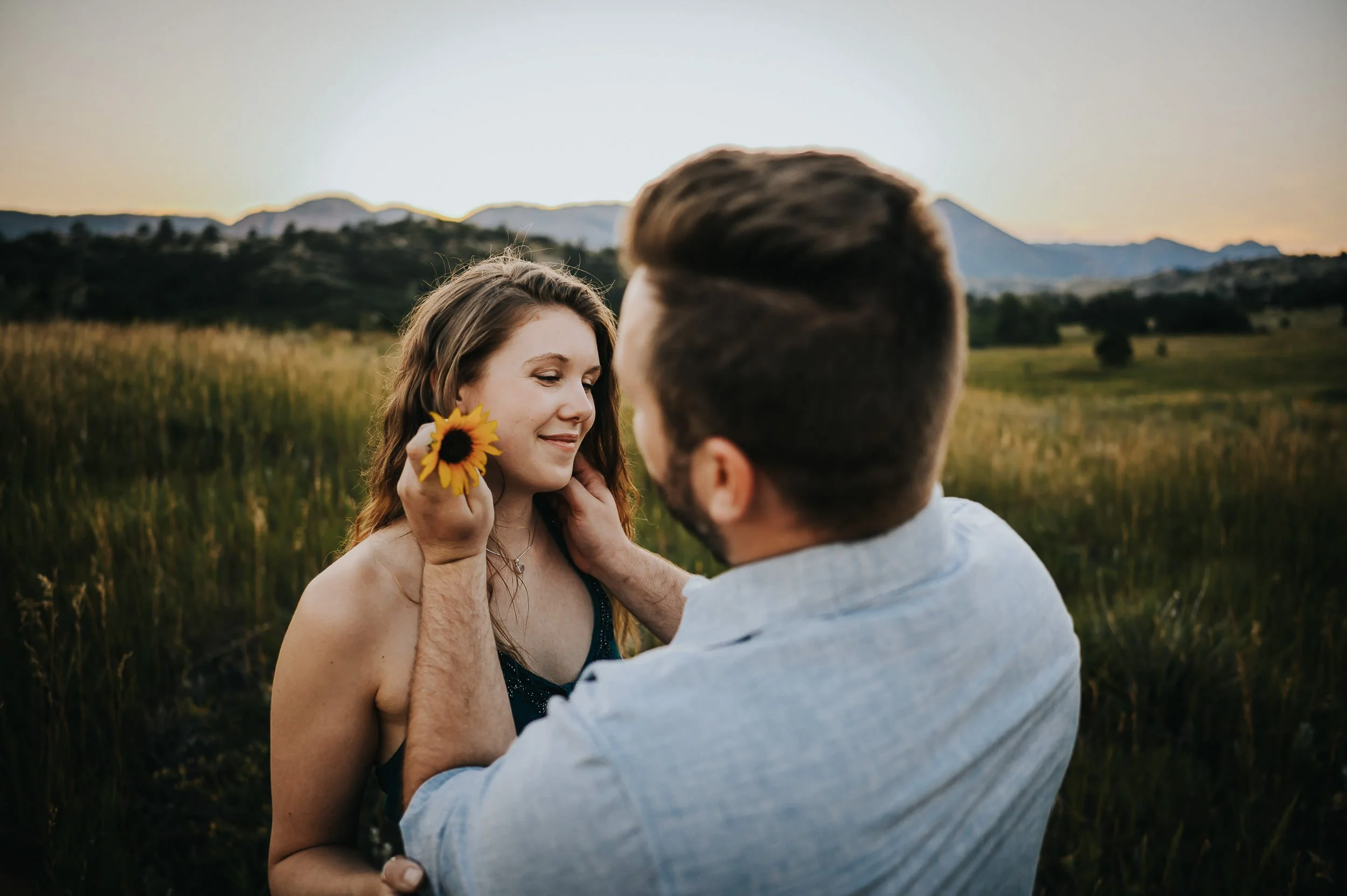 A woman holds a sunflower as her partner leans close from behind, warm and gentle portrait.