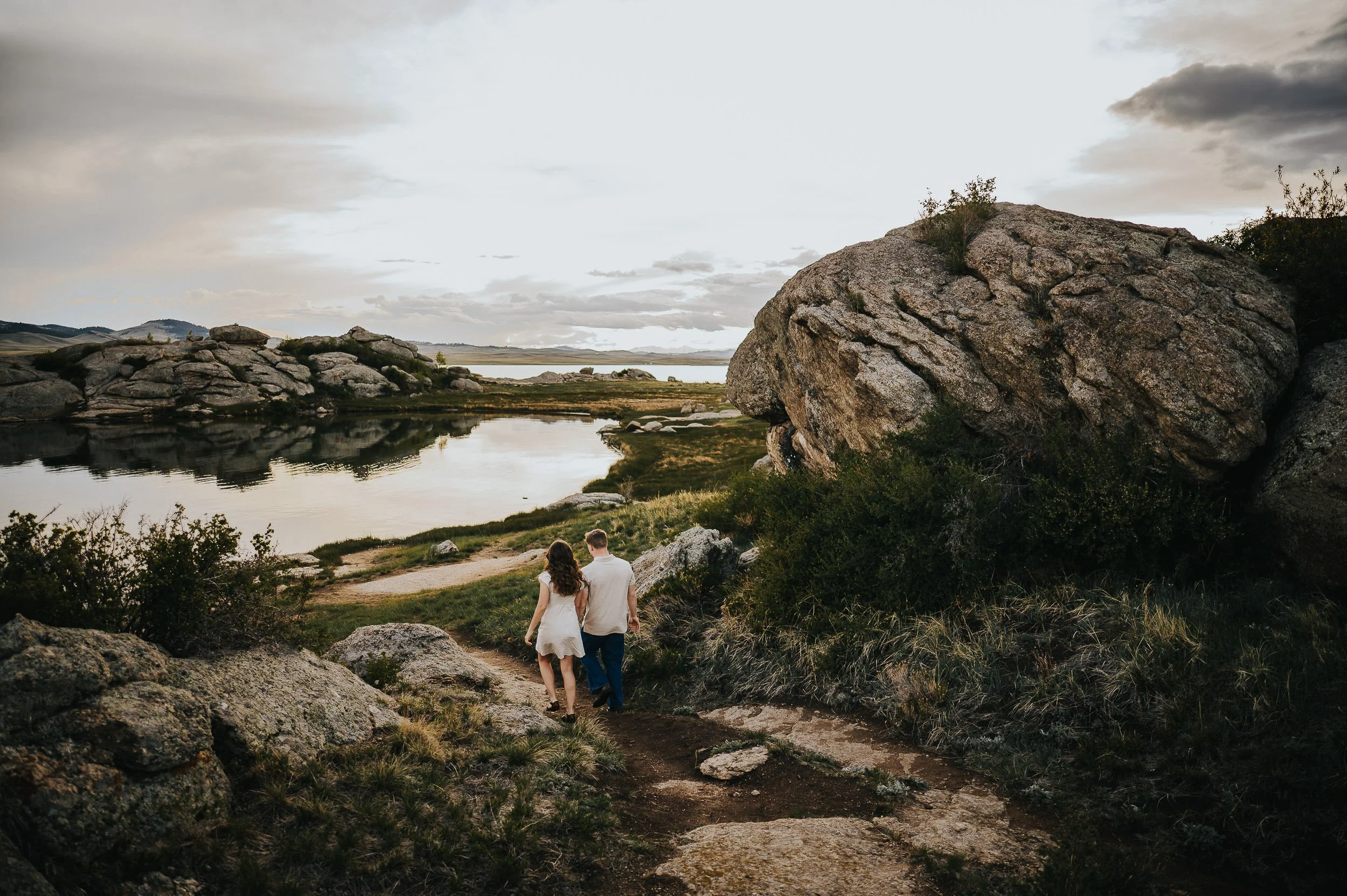 A couple stands small in a wide rocky alpine landscape, mountains and sky dominating the frame.
