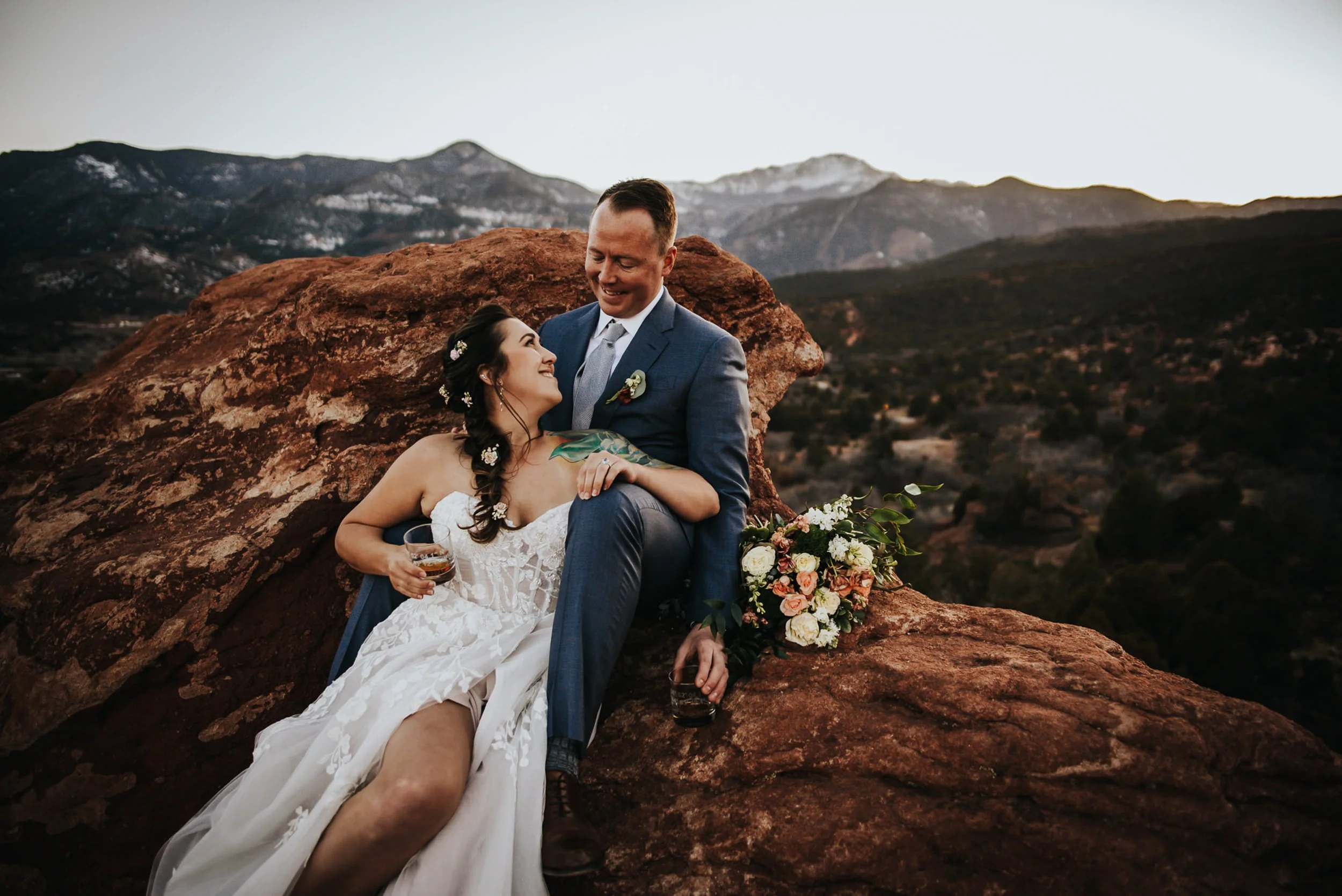 A bride and groom sit together on red rocks, she in a full lace gown with a bouquet, he in a blue suit.
