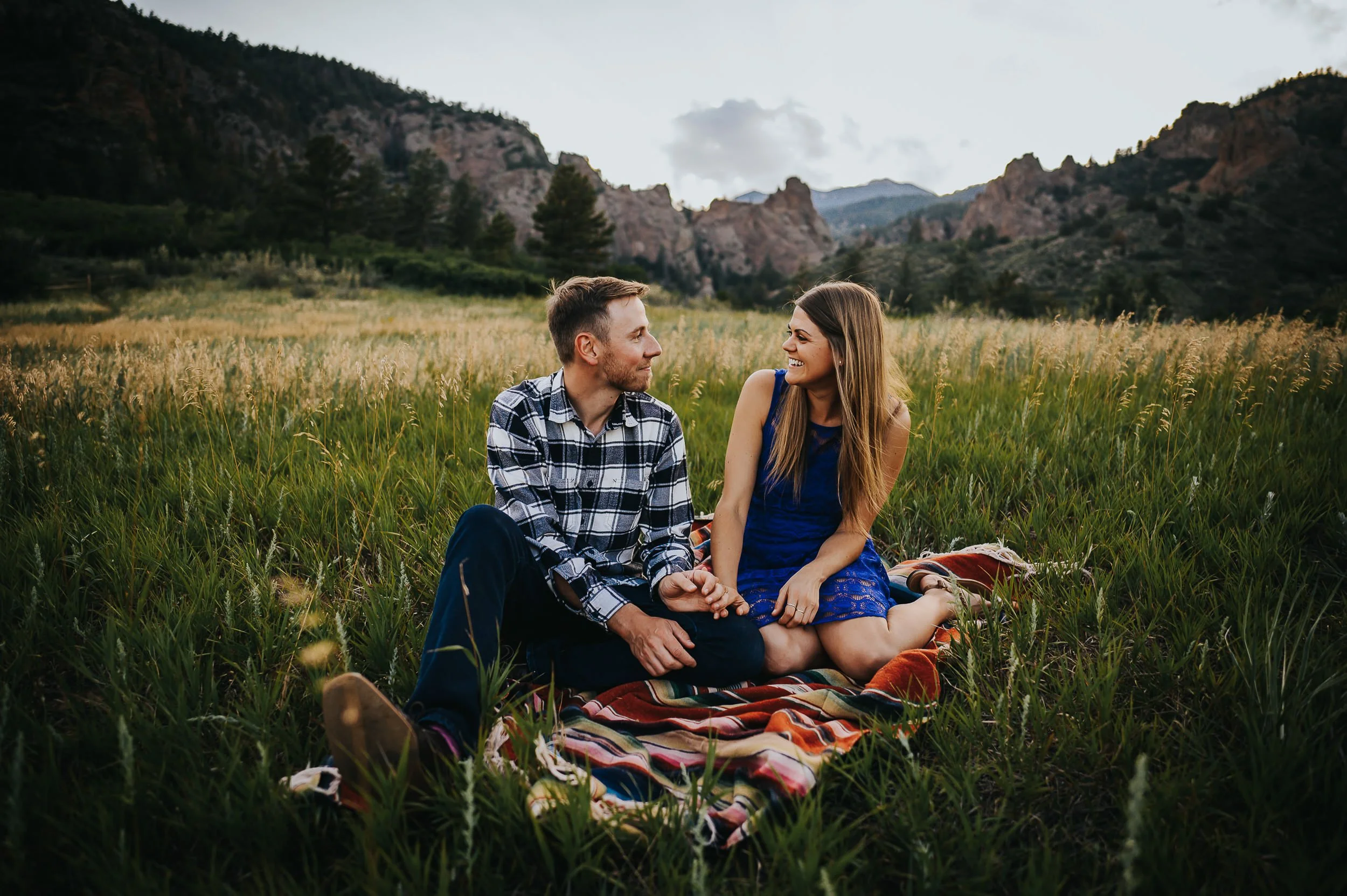 A couple sits together in a lush green Colorado mountain meadow on a blanket, relaxed and laughing.