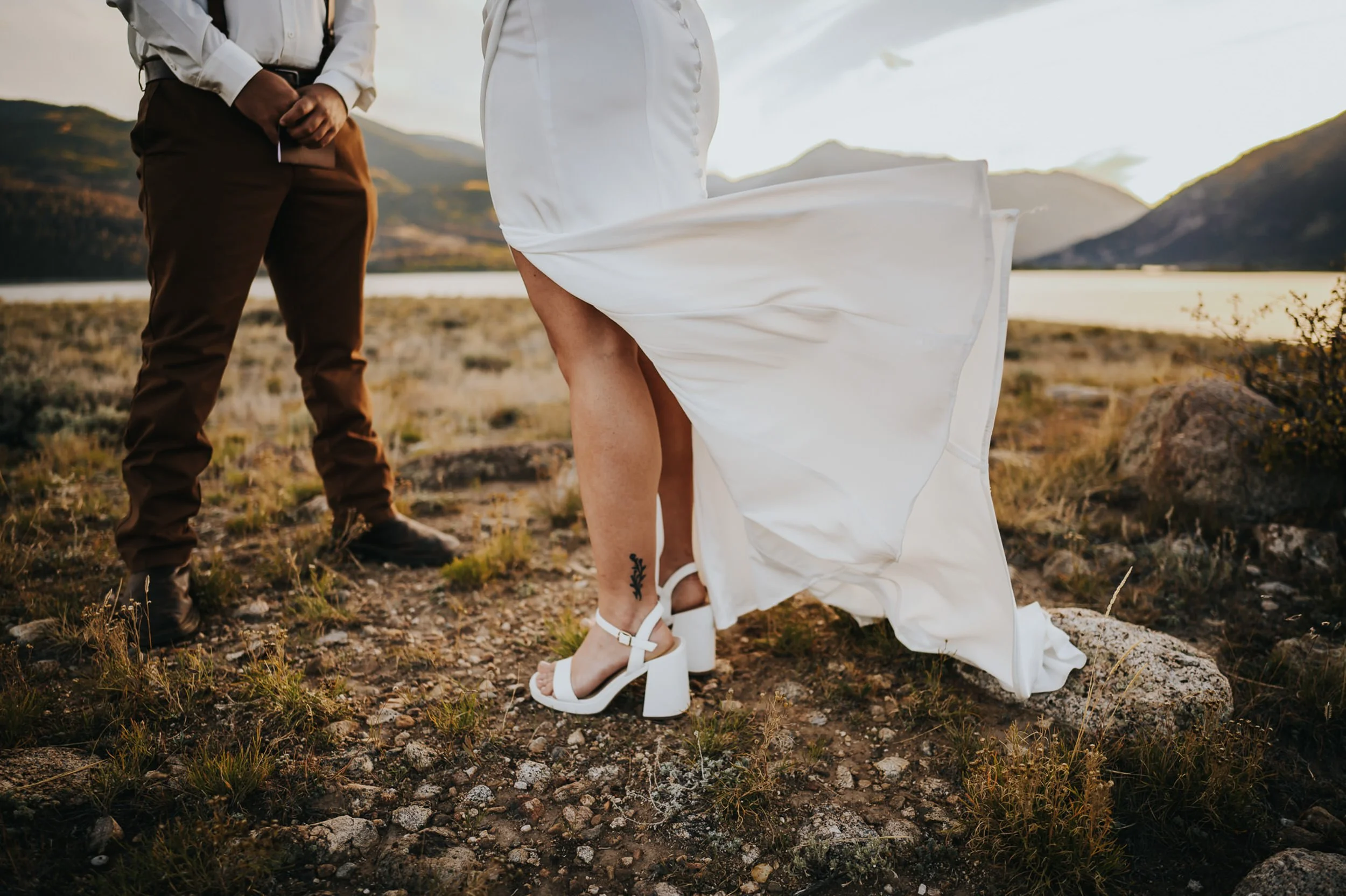 A low angle shot of a bride's white heels and dress hem blowing in the wind, her groom's boots beside her in an open landscape.