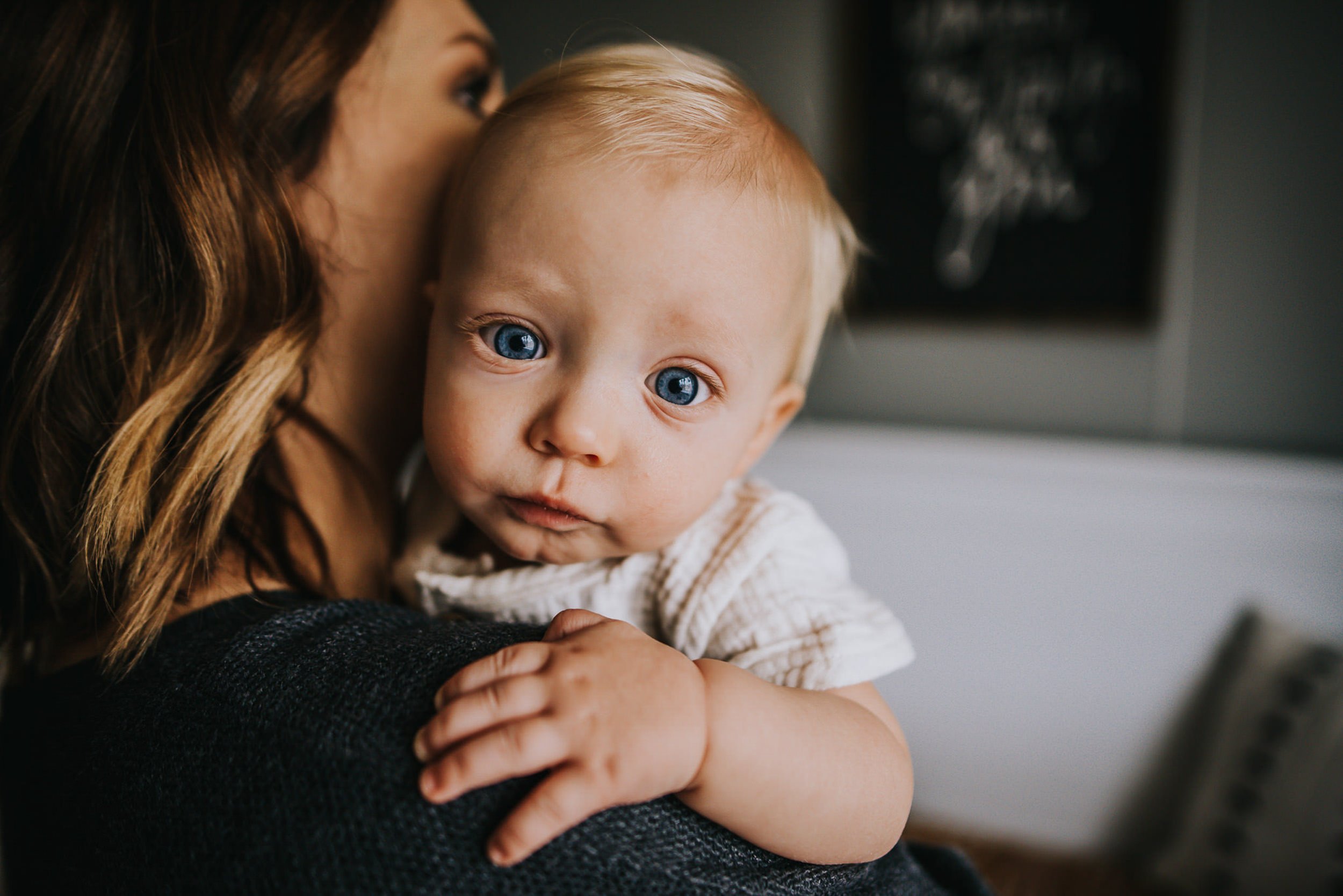 A mother holds her blue-eyed baby close in a warm indoor setting, baby looking directly at camera.