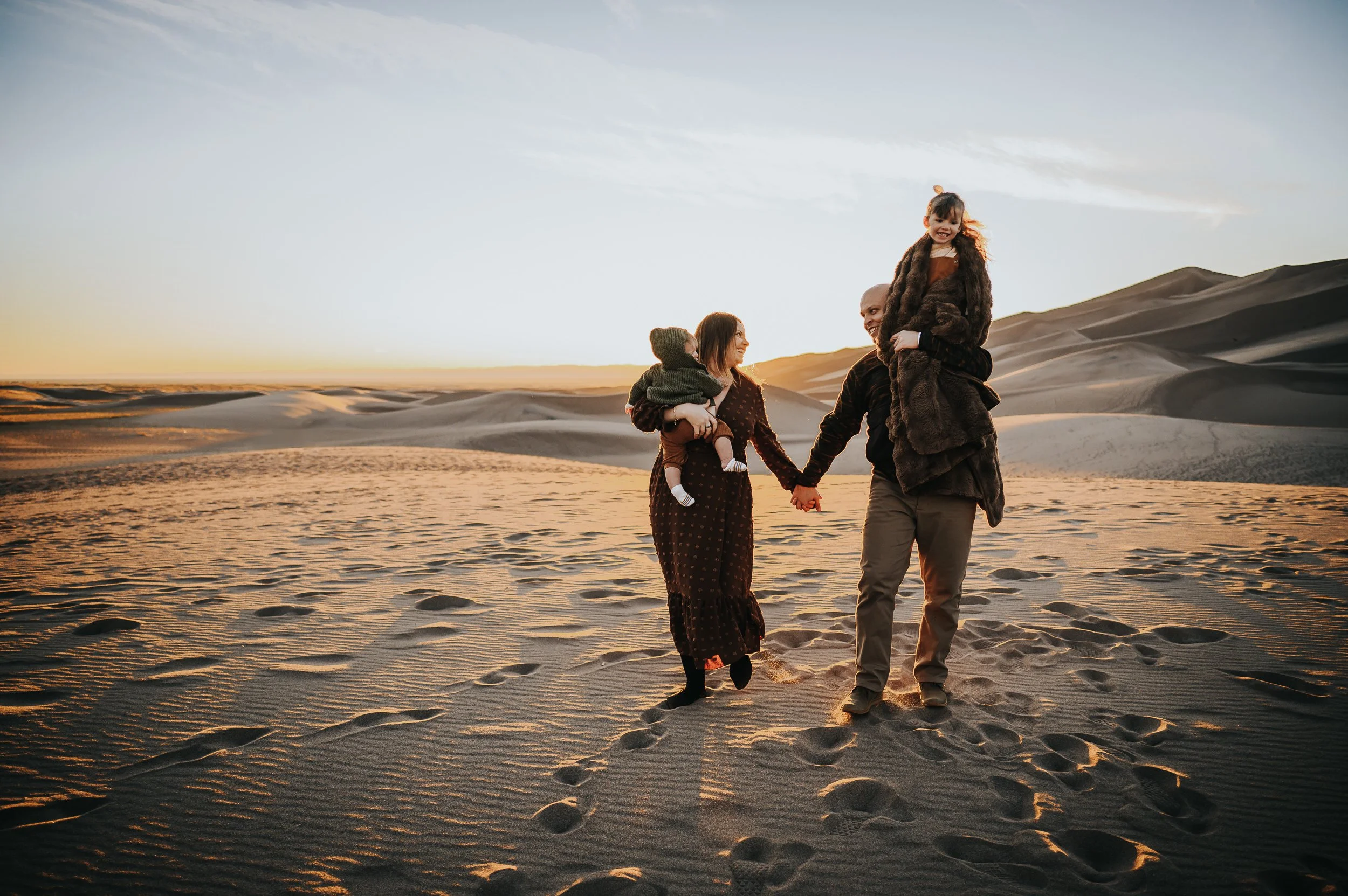 Family of four holding hands walking across the Great Sand Dunes in Colorado at sunset.