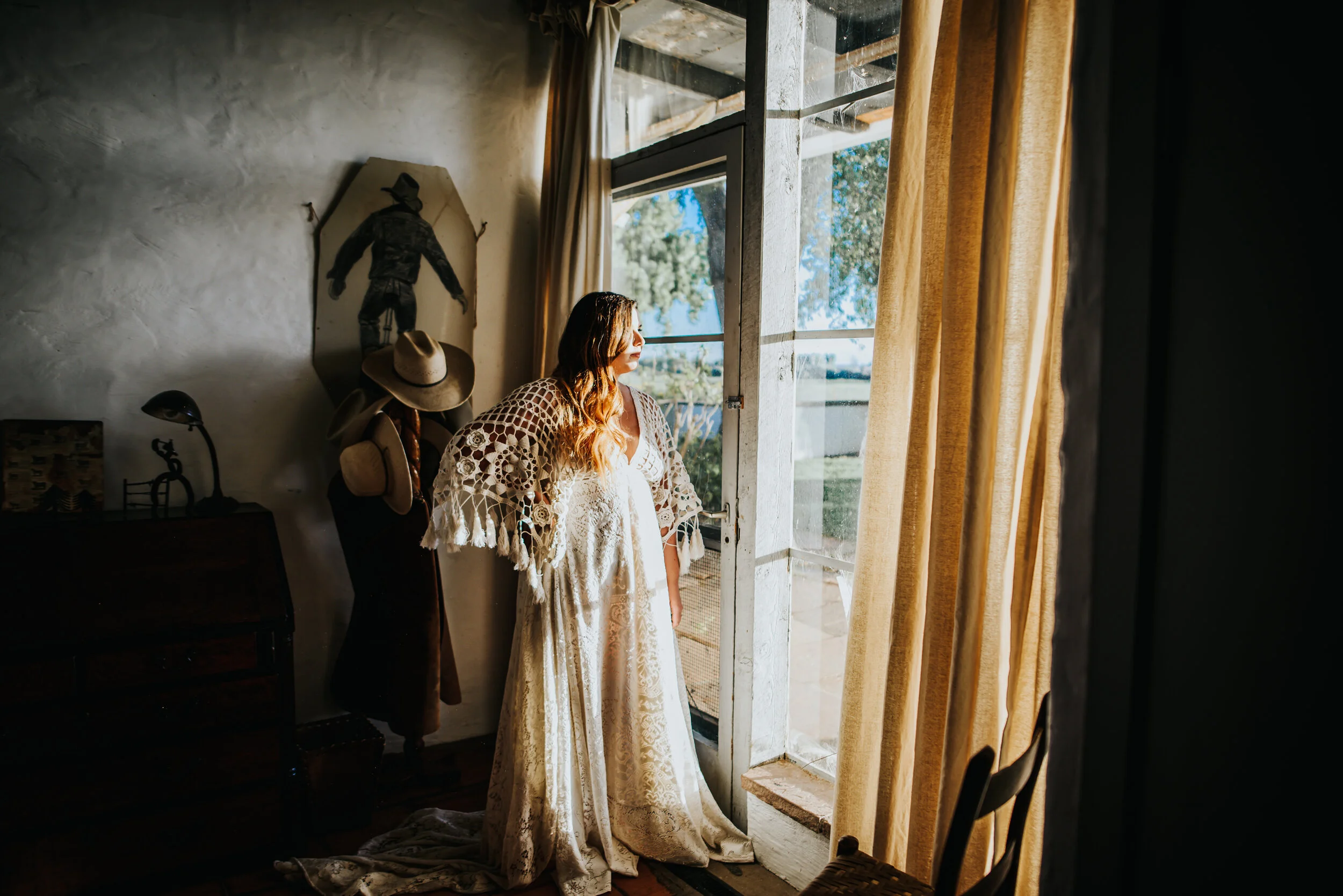 Bride in a wedding gown standing beside tall curtained windows in a warmly lit room.