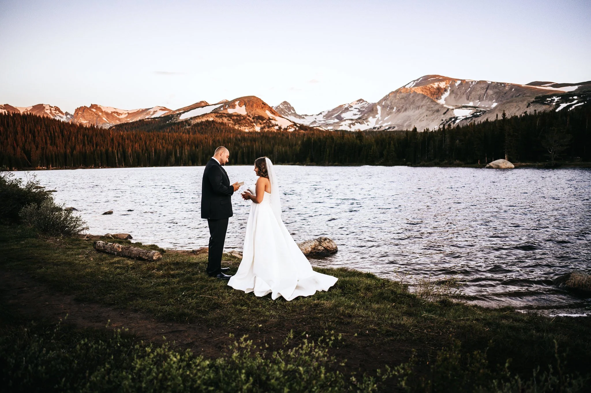Couple standing face to face exchanging vows beside an alpine lake with snow-capped mountains behind them.