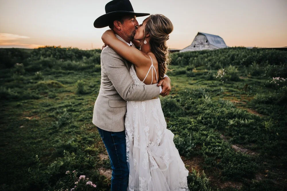 Couple sharing a kiss in a lush green field at a Nebraska Sandhills ranch wedding, groom wearing a cowboy hat.