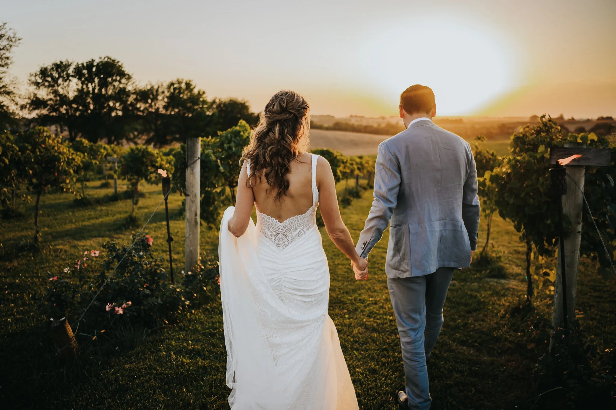 Bride and groom walking hand in hand through a vineyard at golden hour, bride in a lace gown.