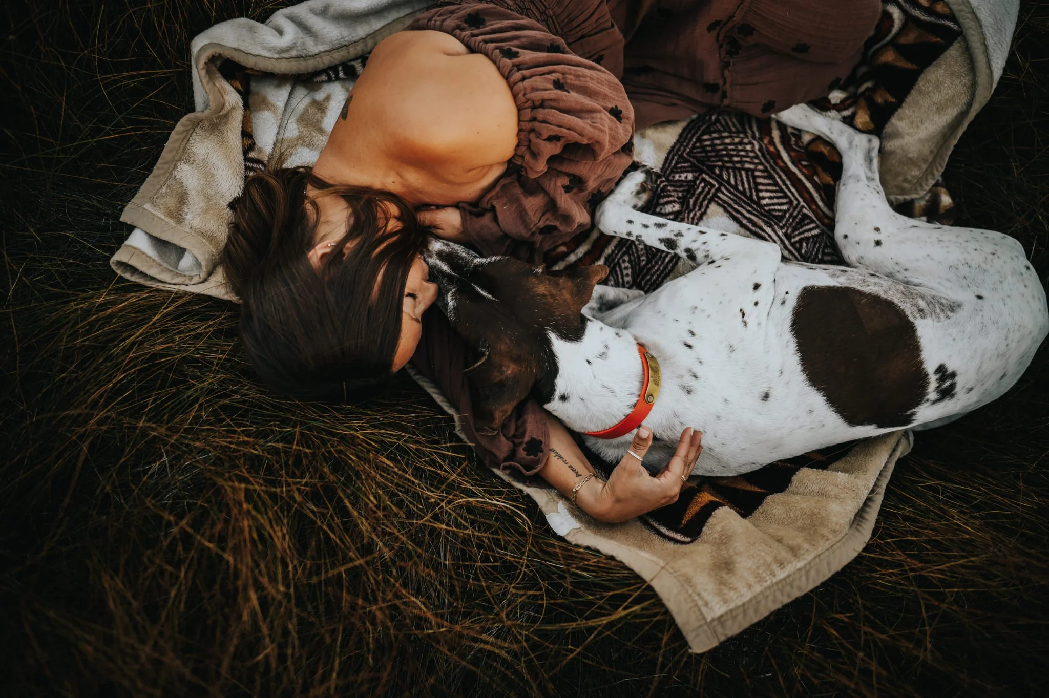 Woman lying in grass with her spotted dog surrounded by layered blankets, showcasing texture in family photography.