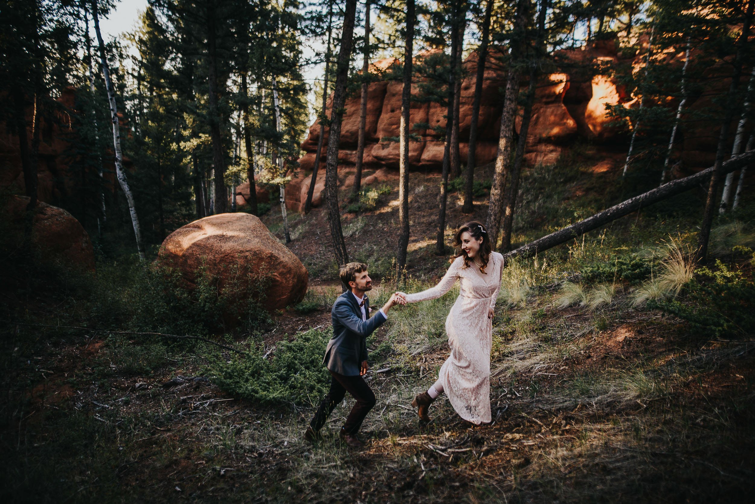 Bride and groom running and laughing together through a pine forest at golden hour.