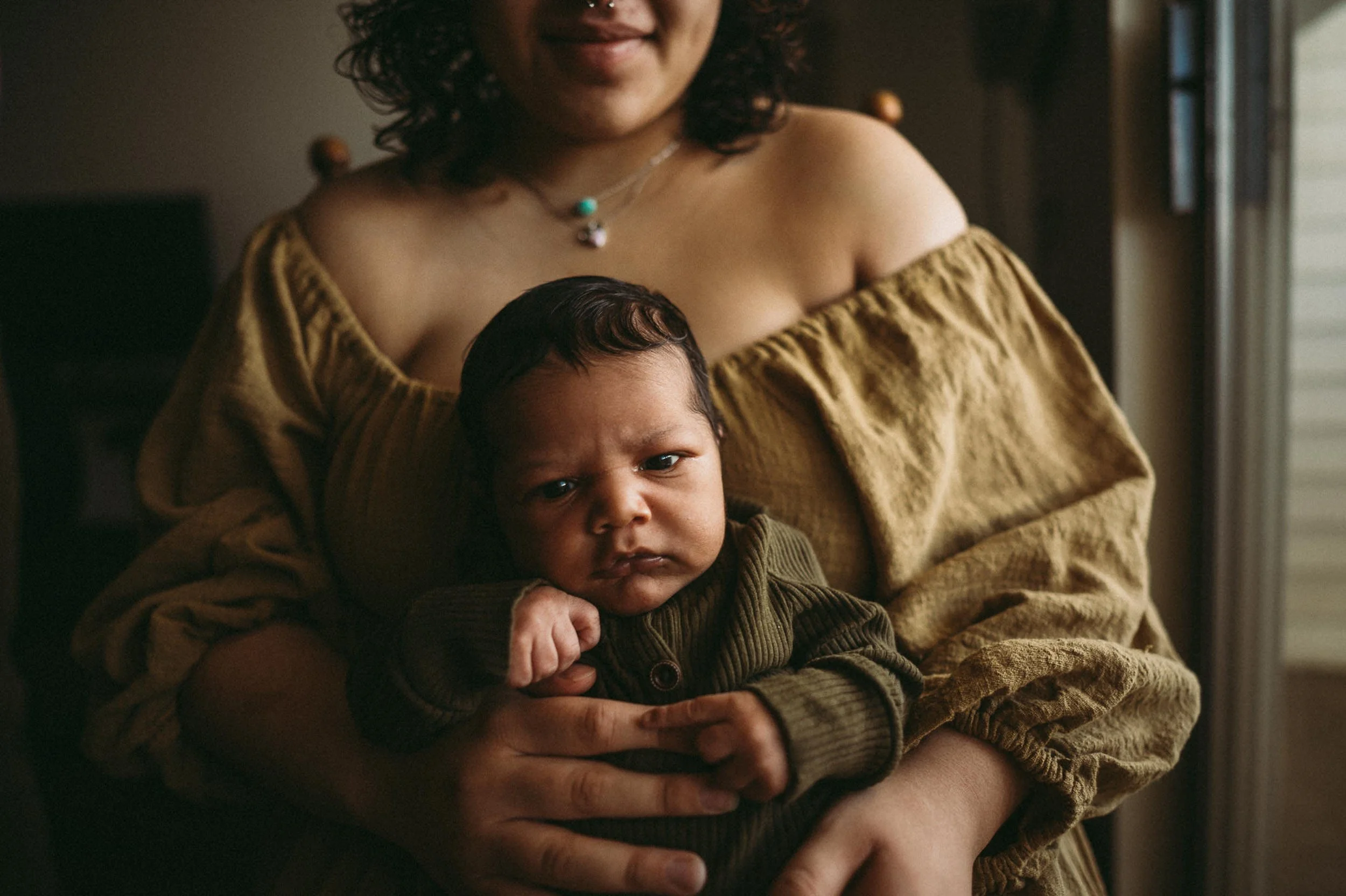 mother cradles her newborn wrapped in gold, soft indoor light falling over them both.