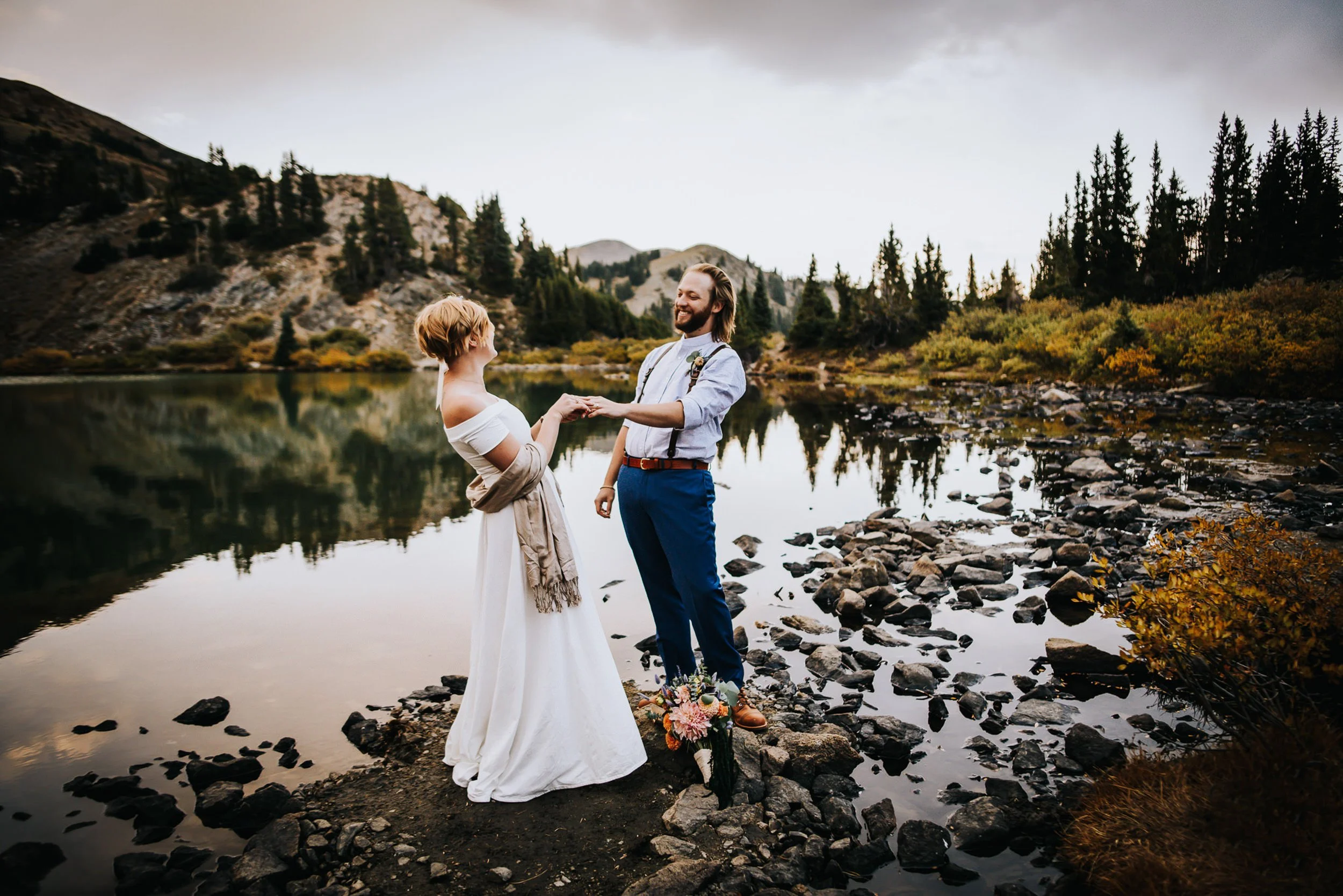 A bride and groom stand at the edge of a reflective alpine lake, surrounded by rocky Colorado wilderness.