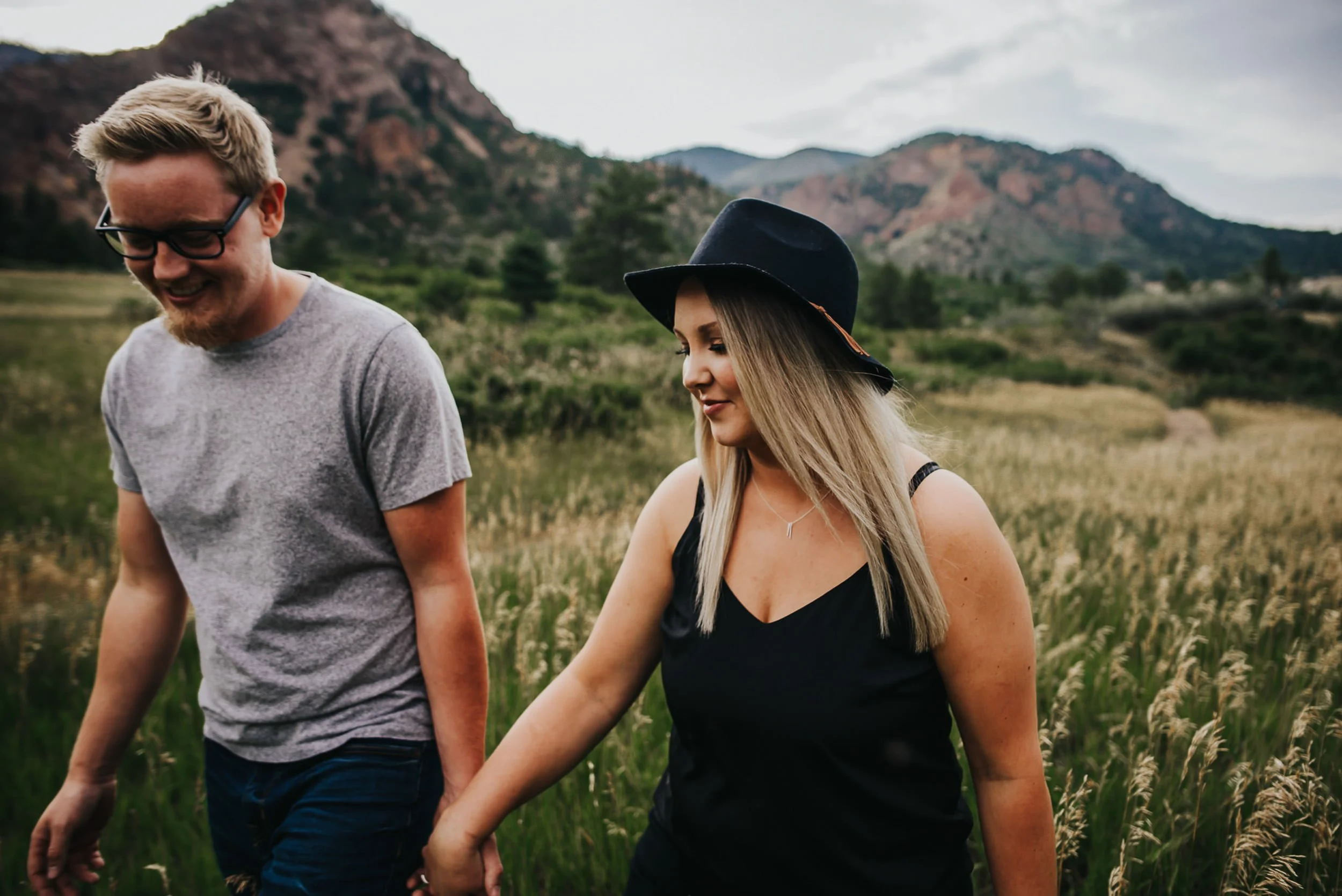 A couple walks hand in hand through a Colorado foothills meadow, she in a black hat, red rock formations behind them.