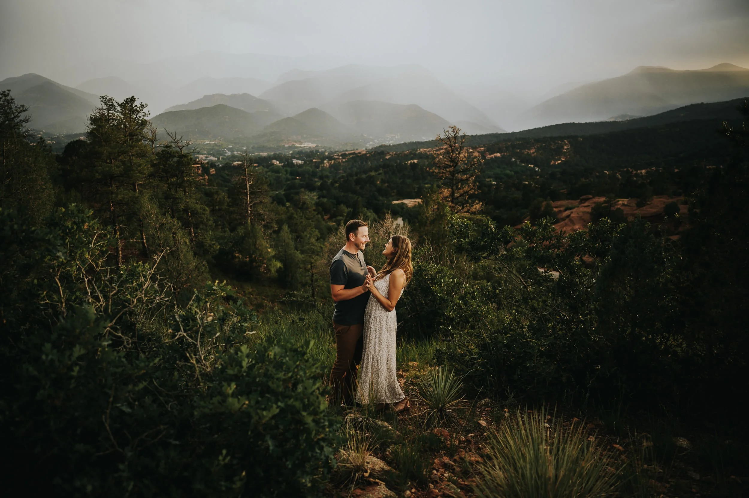 A couple stands together in a misty Colorado mountain landscape, wide shot with peaks behind them.