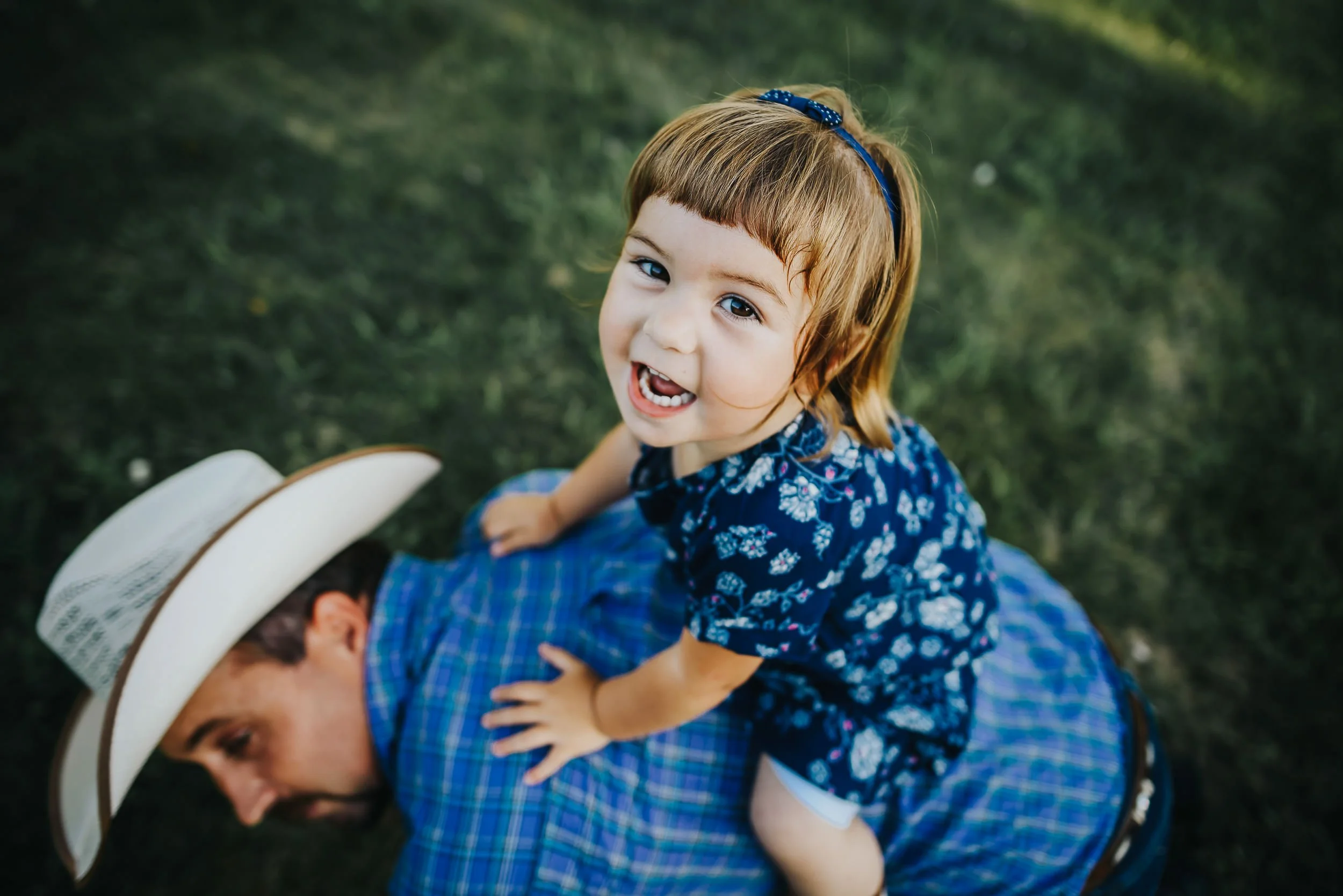 A little girl dressed in blue rides on the back of her dad in the grass, laughing.
