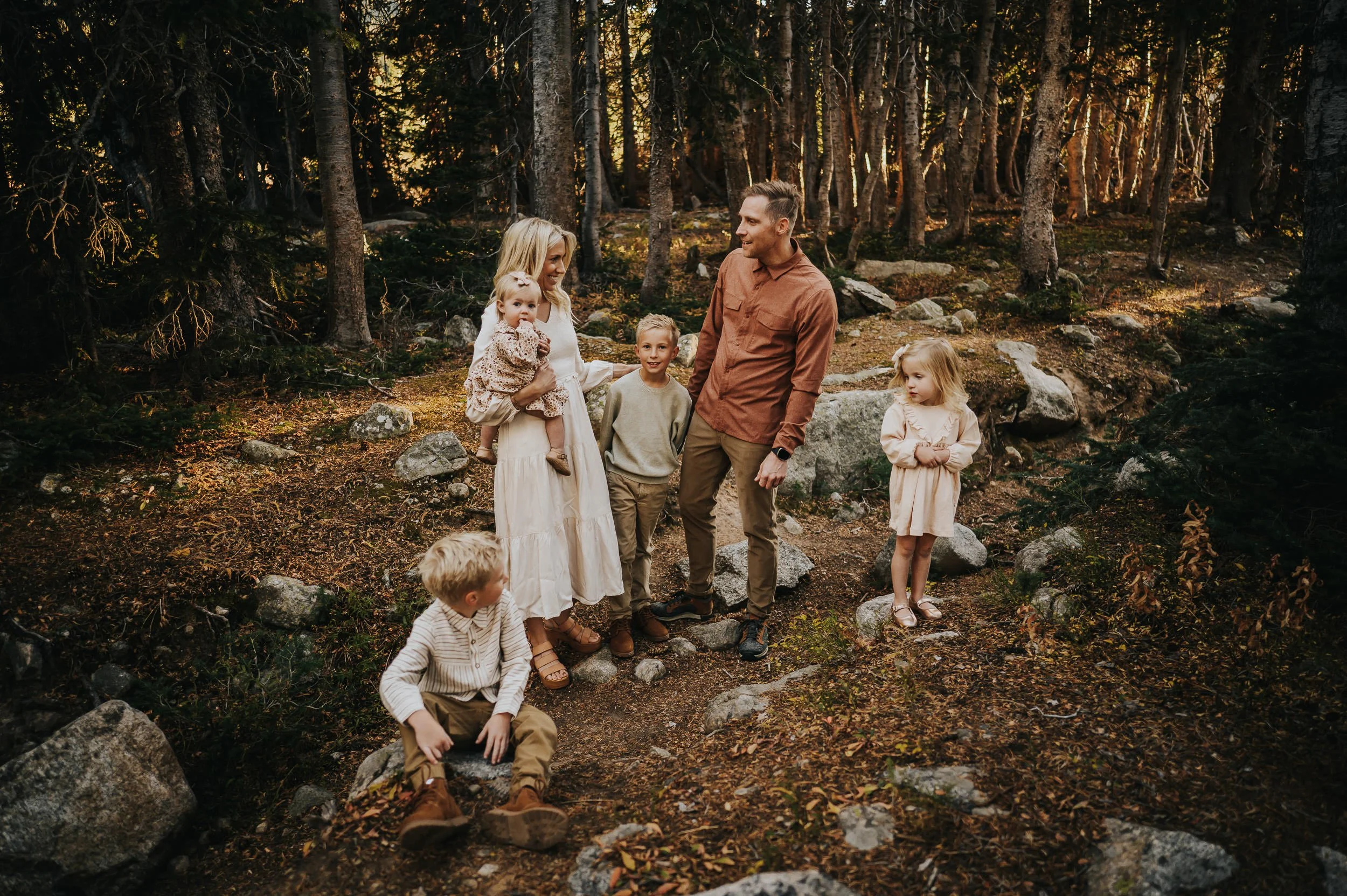 Family of six standing in a Colorado evergreen forest, coordinated in cream, rust, and dusty rose tones with tall pine trees behind them.