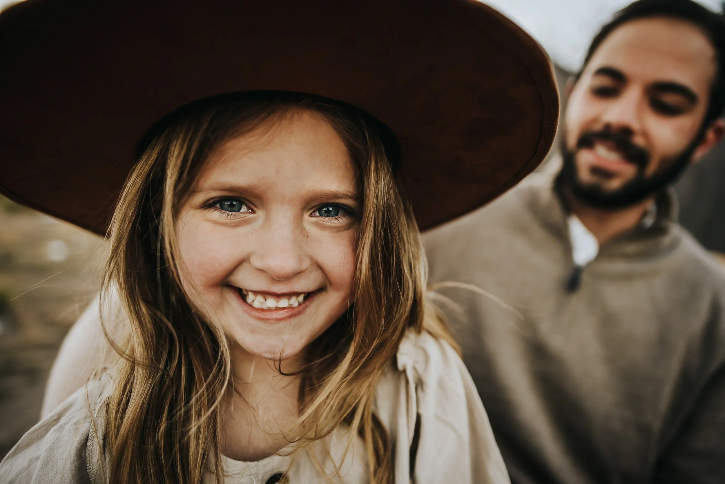A young girl with bright blue eyes smiles wide beneath a wide-brim hat, her dad just visible behind her.
