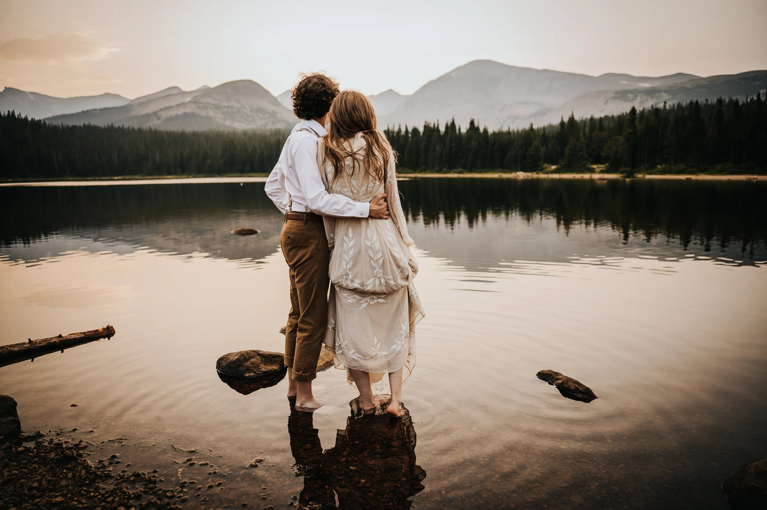 Couple standing together at the edge of a still mountain lake surrounded by pine trees.