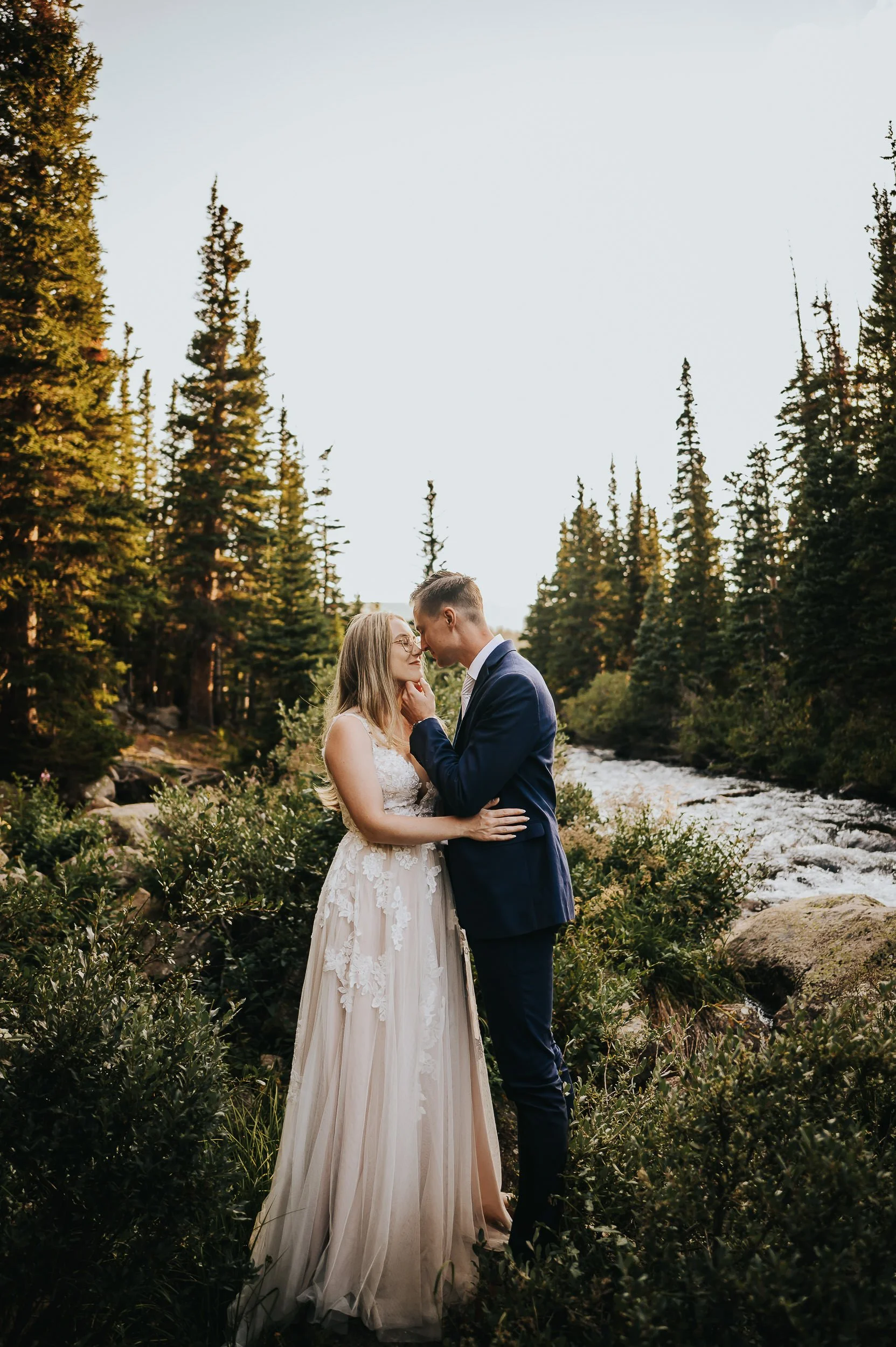 A bride and groom stand together in a Colorado alpine meadow with rocky peaks and pine forest behind them.