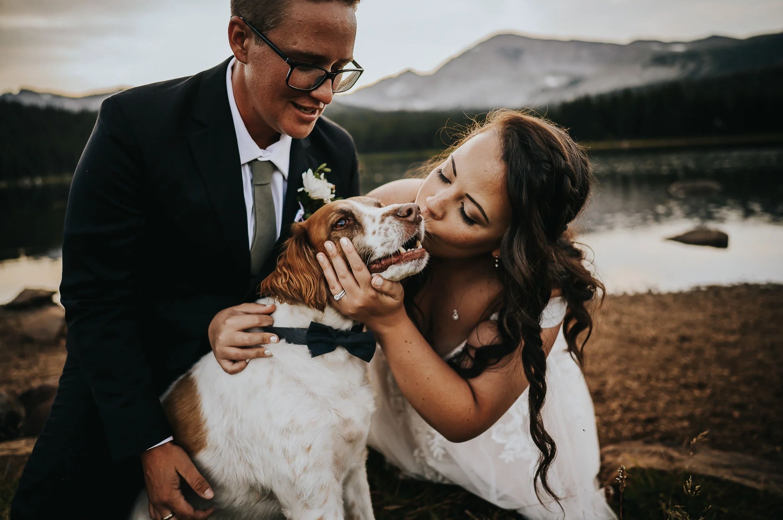 Bride and groom laughing together beside a mountain lake while their dog joins in.