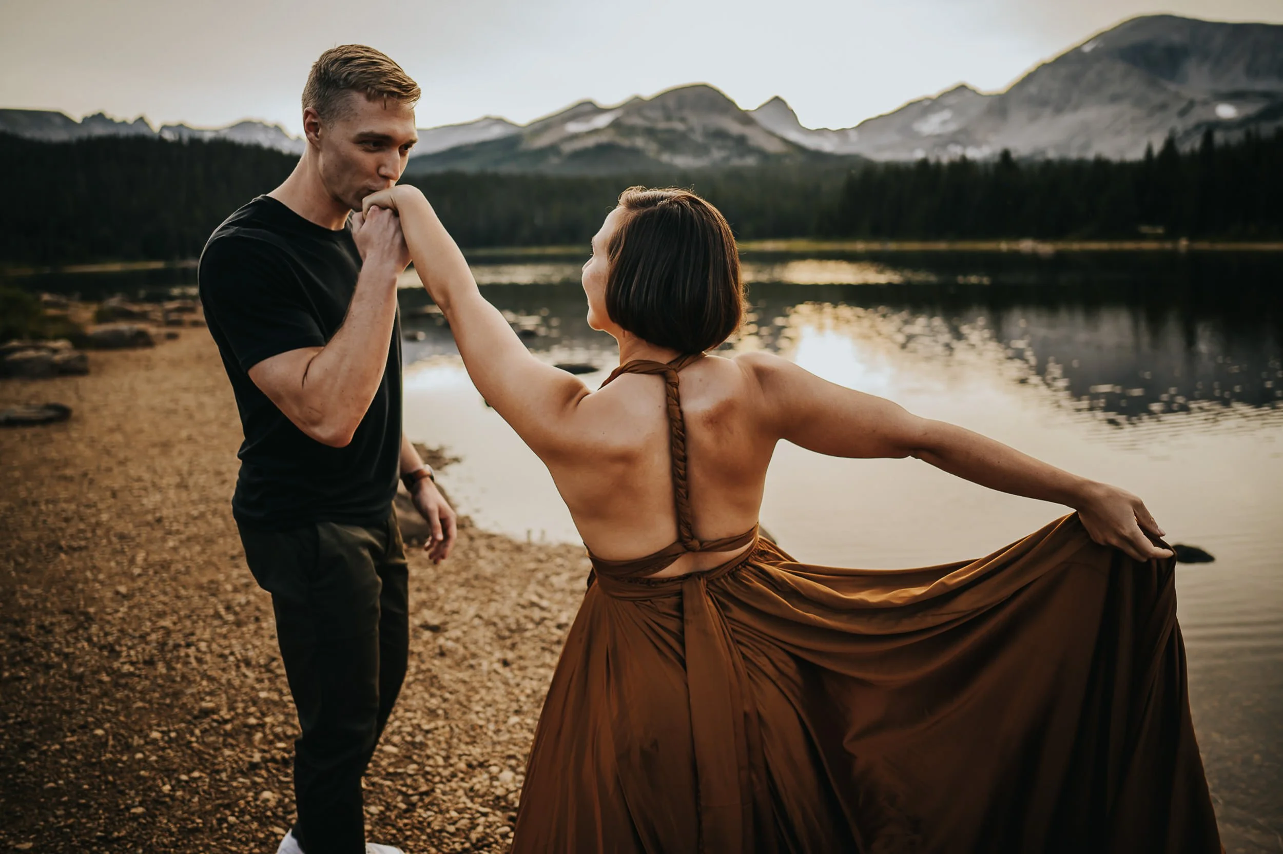 A groom spins his bride by a mountain lake, her brown skirt flaring out, peaks behind them.