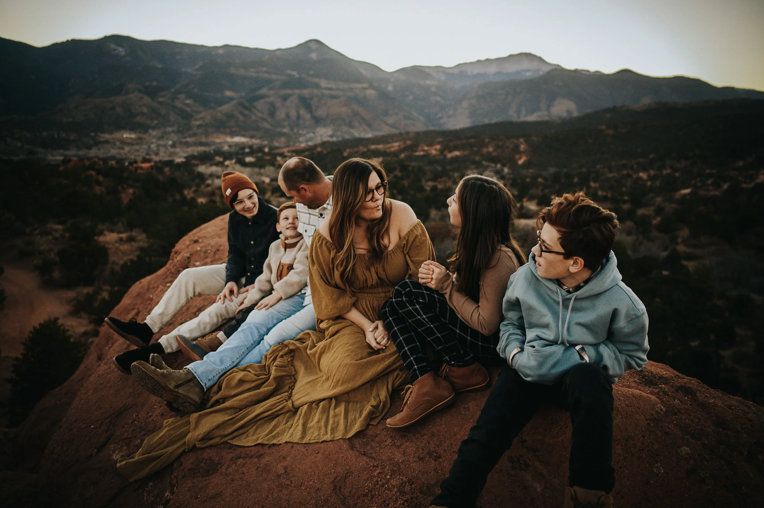 Family of six sitting together on red rock formations at Garden of the Gods with Pikes Peak and mountains behind them.