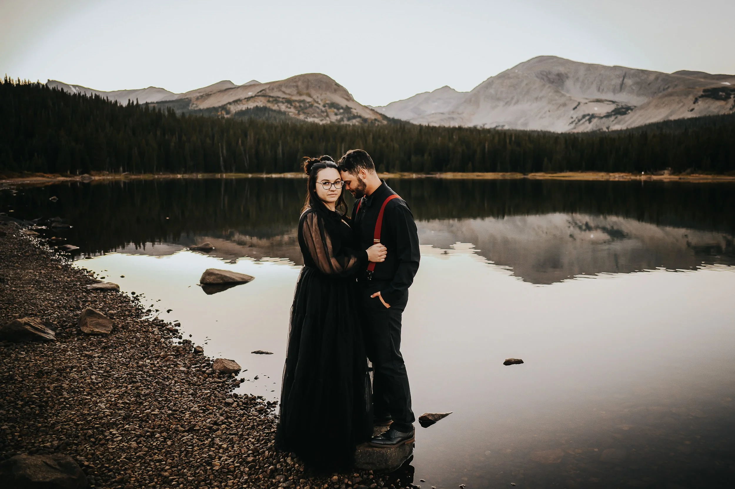 A couple stands close together at the edge of a reflective Colorado mountain lake at dusk.