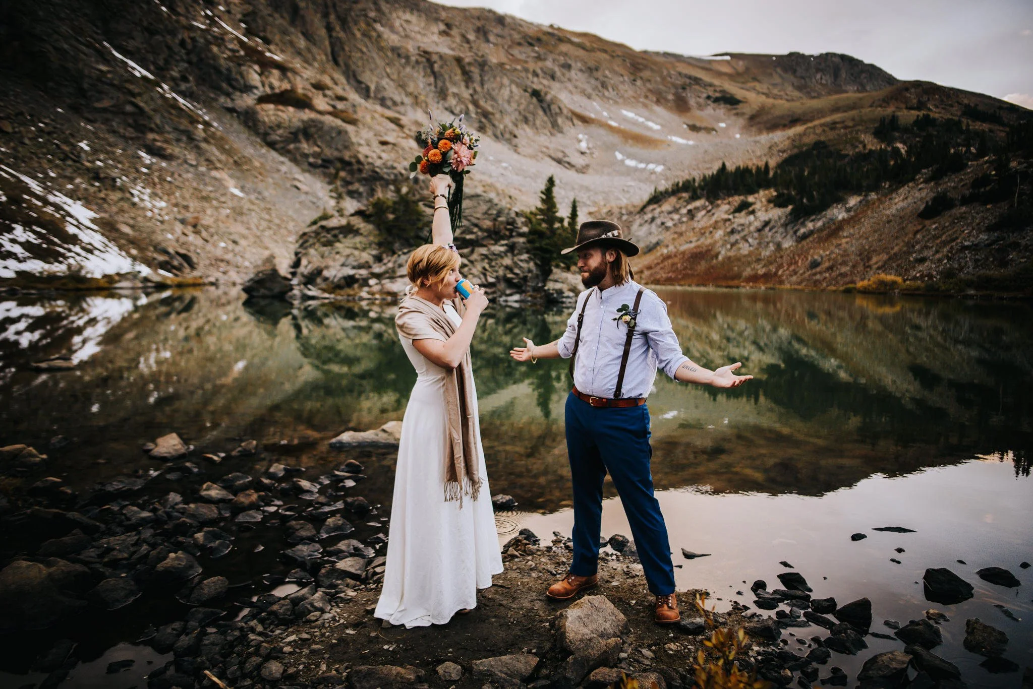 Couple joyfully celebrating beside an alpine lake, woman raising her bouquet in the air with mountains behind them.