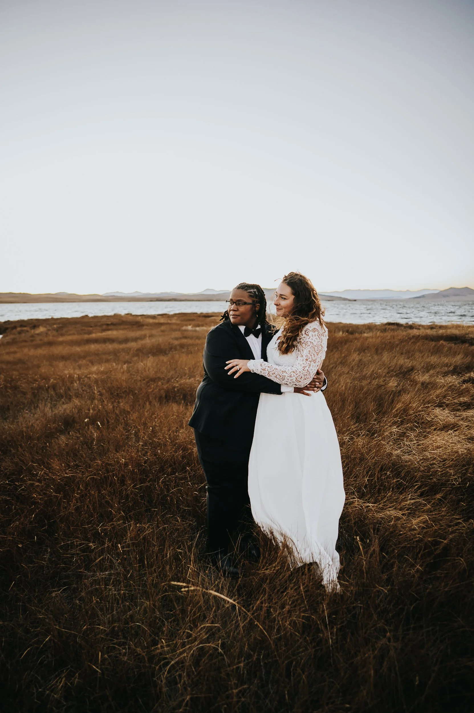 Couple holding each other in a wide open field under a soft overcast sky, bride in a white dress.