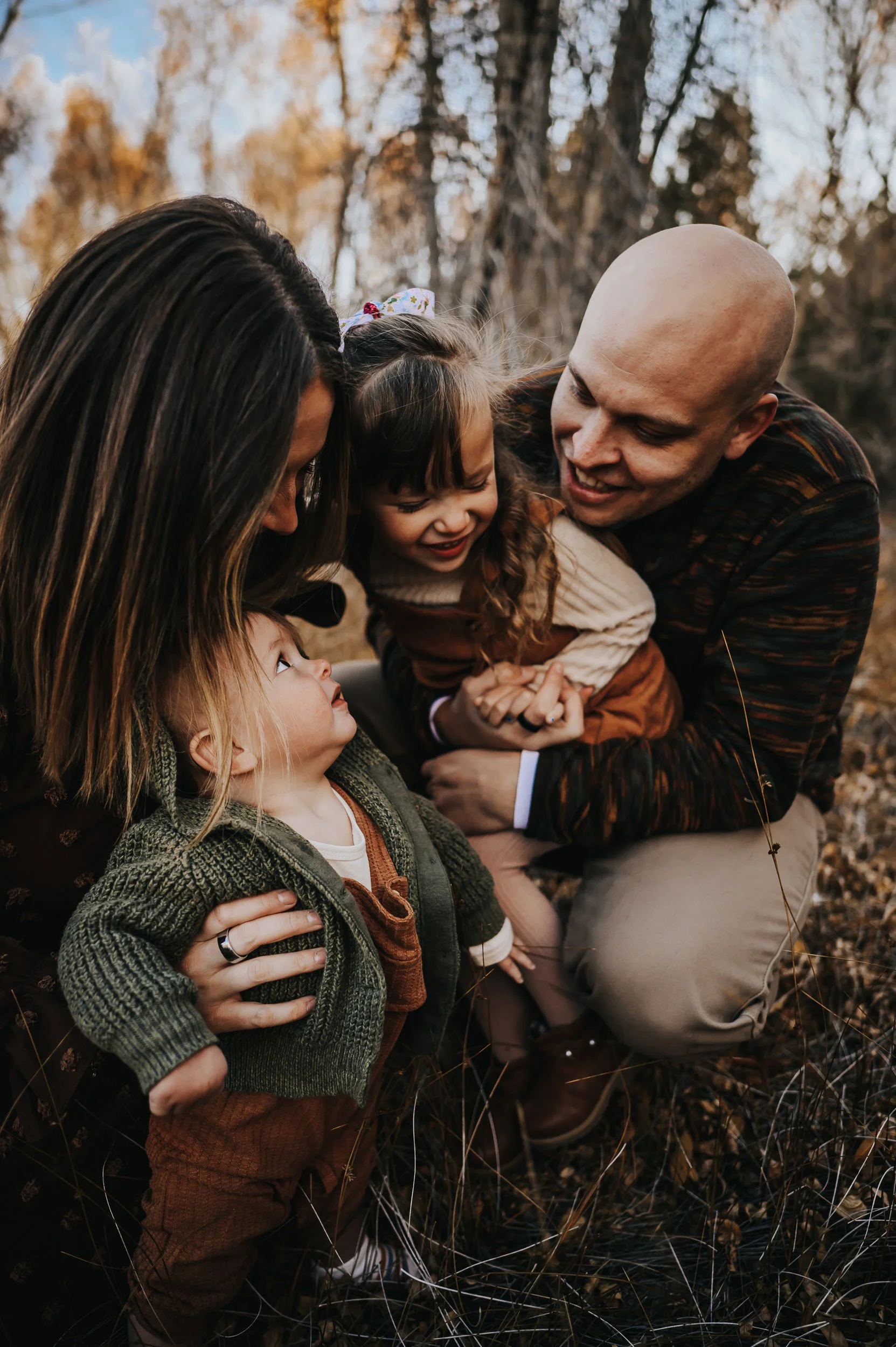 Family of four in intimate fall portrait with autumn trees behind them, coordinated in olive, rust, and cream earth tones.
