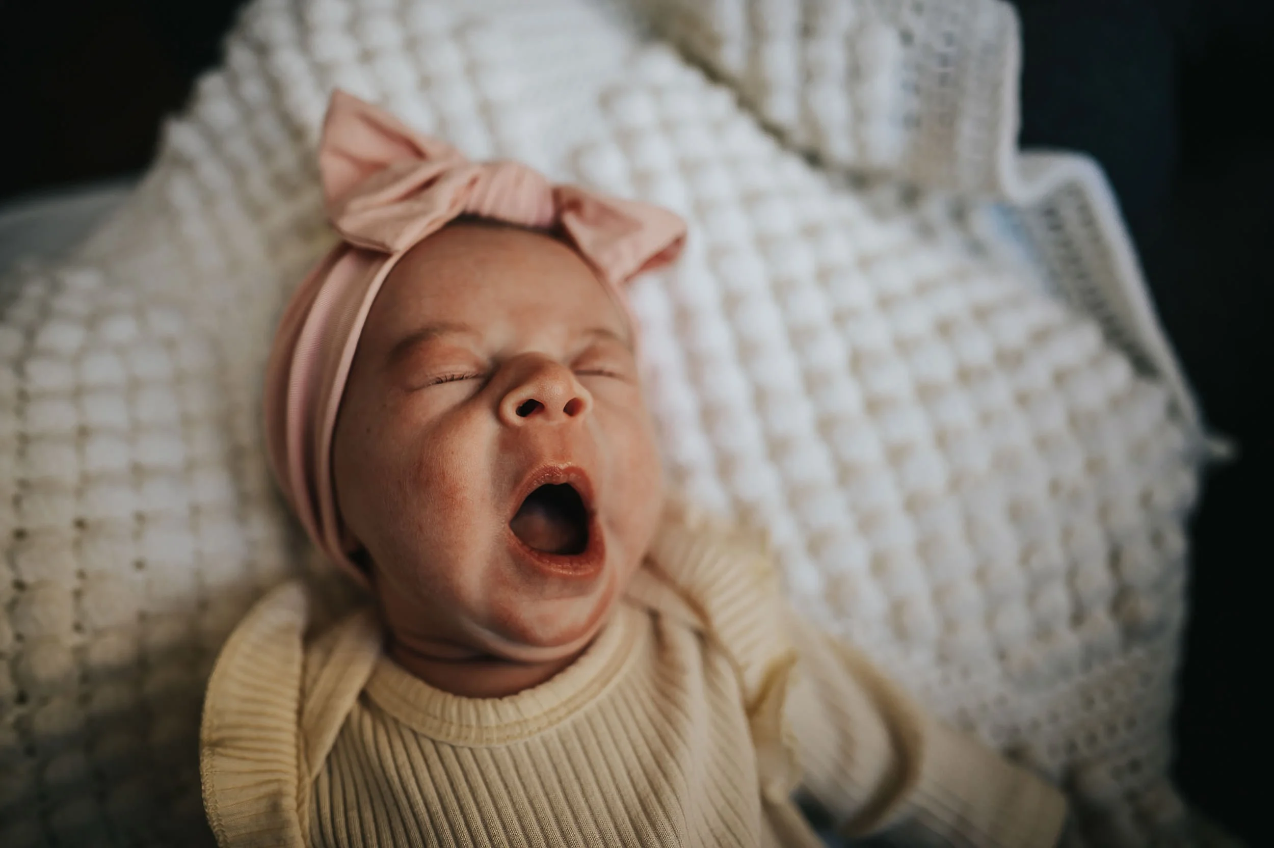 Newborn baby yawning while wearing a cream outfit with a pink bow.