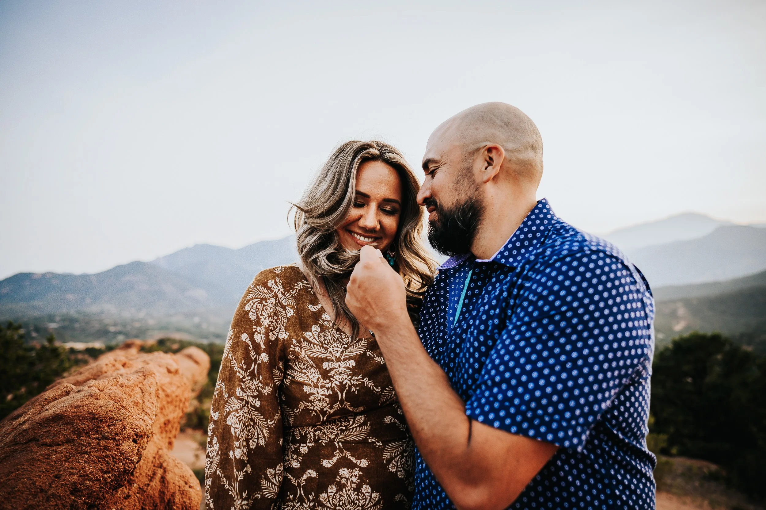 A bearded man tenderly touches his partner's face, she looks up at him laughing, Colorado mountains behind them.