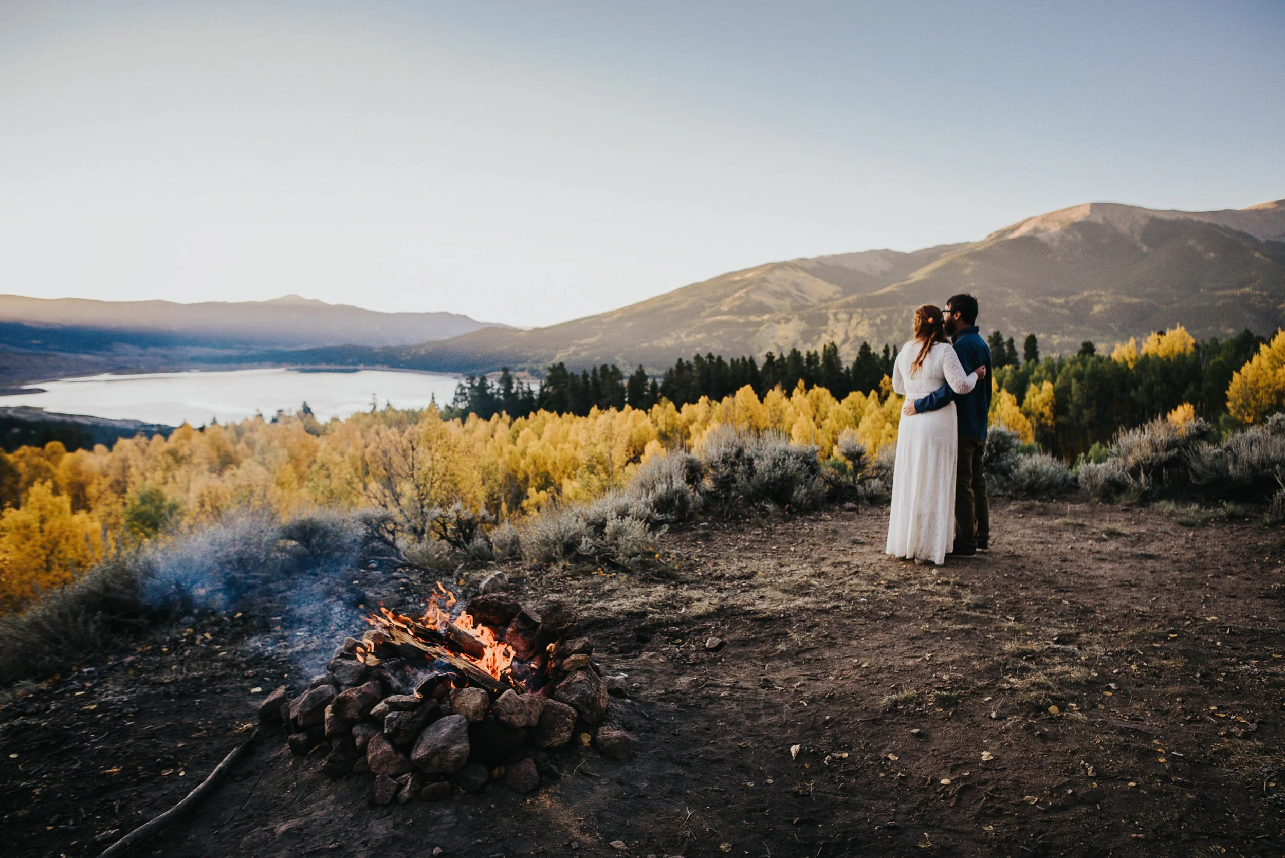 Couple standing near a campfire surrounded by golden aspen trees beside a mountain lake at sunrise.