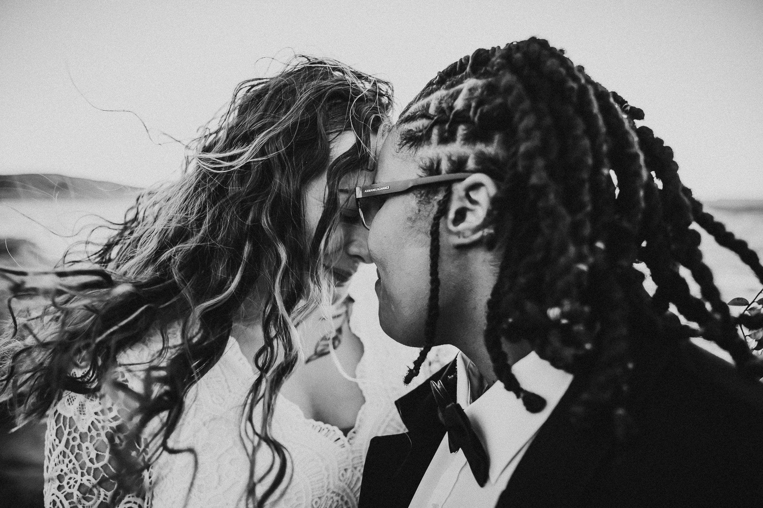 A couple shares a close kiss in black and white, her locs blowing across their faces.