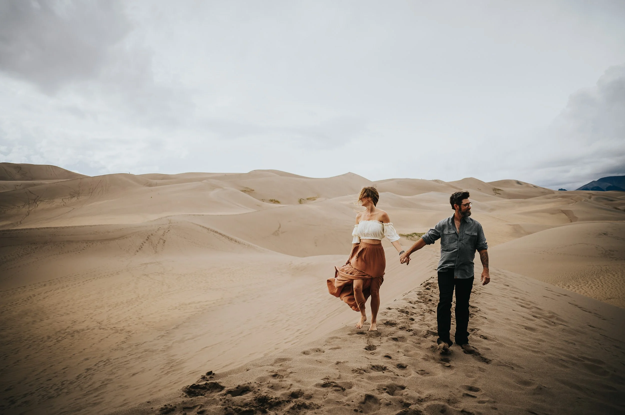 A couple walks hand in hand through the Great Sand Dunes, wide open desert landscape around them.