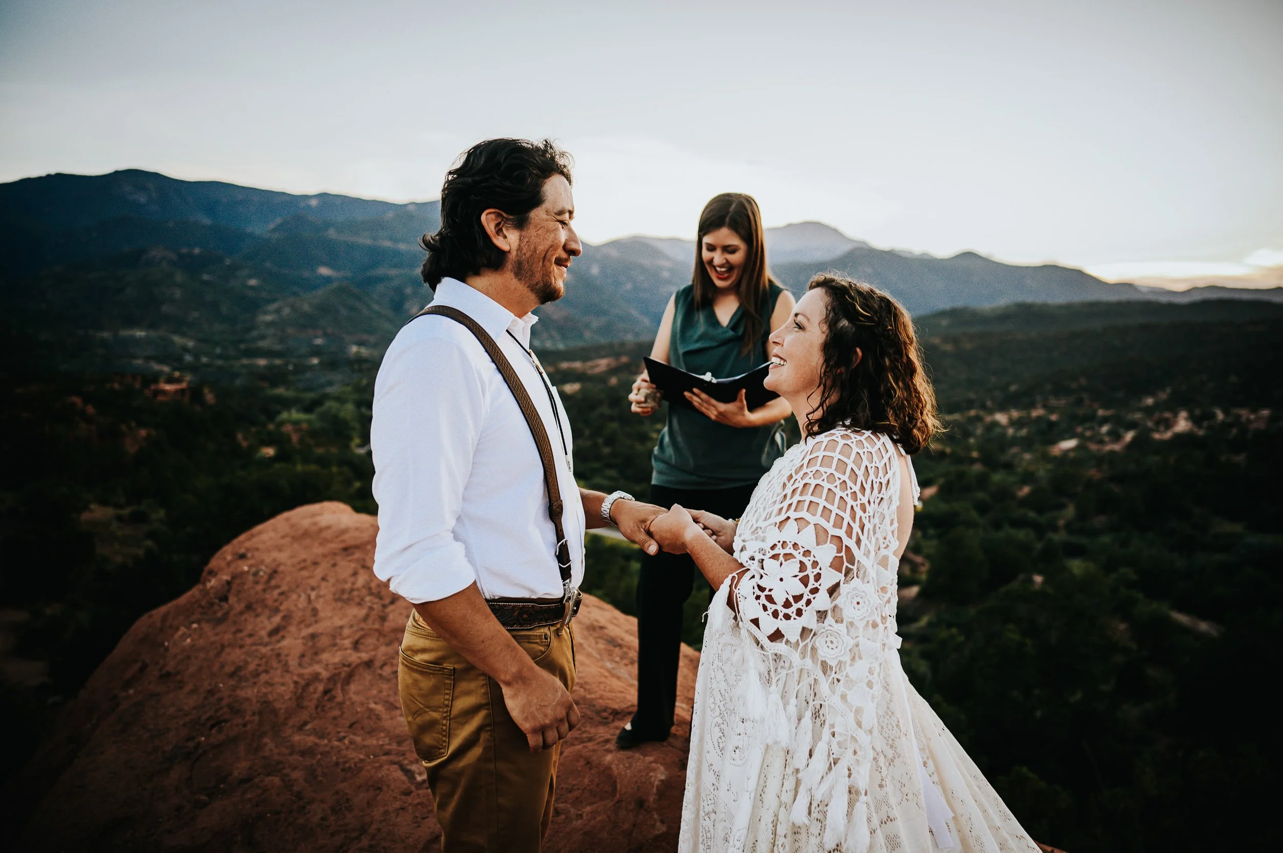 A couple laughs together on red rocks, he in white with suspenders, she in an intricate lace dress.