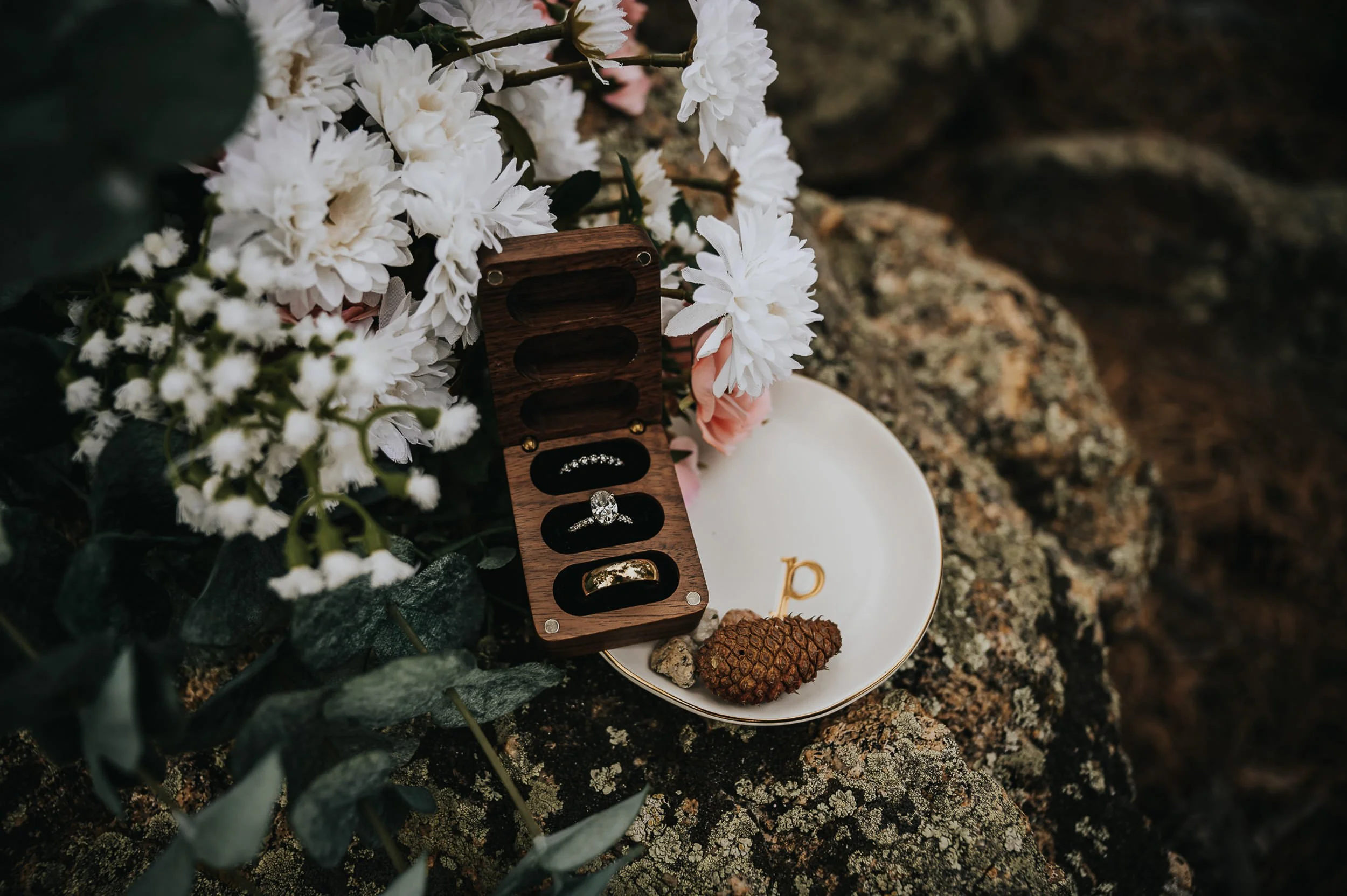 A wedding ring box with two rings rests on a rock beside white daisy florals, detail flatlay.