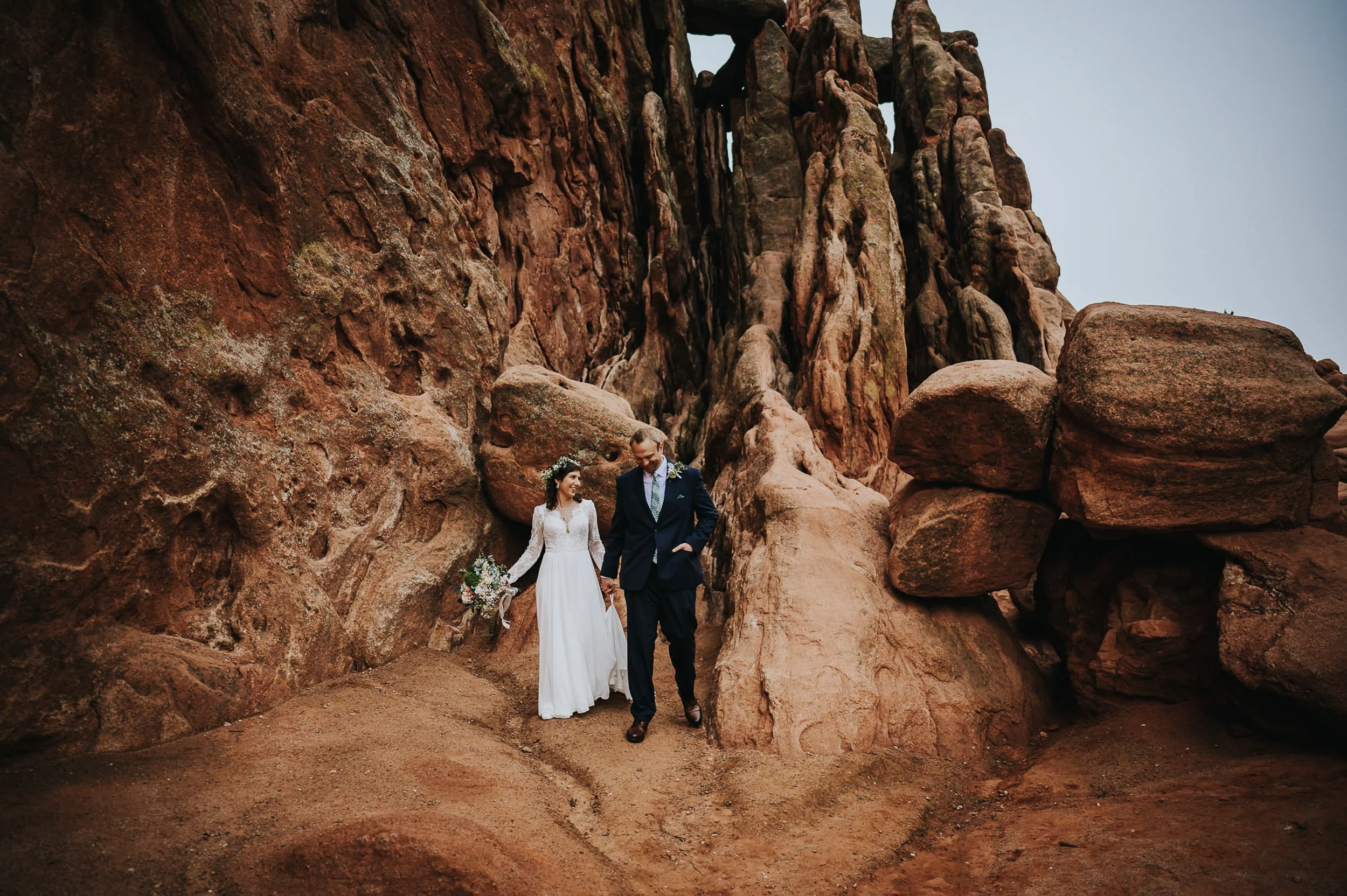 A bride and groom stand together dwarfed by towering red rock walls at Garden of the Gods, wide shot.