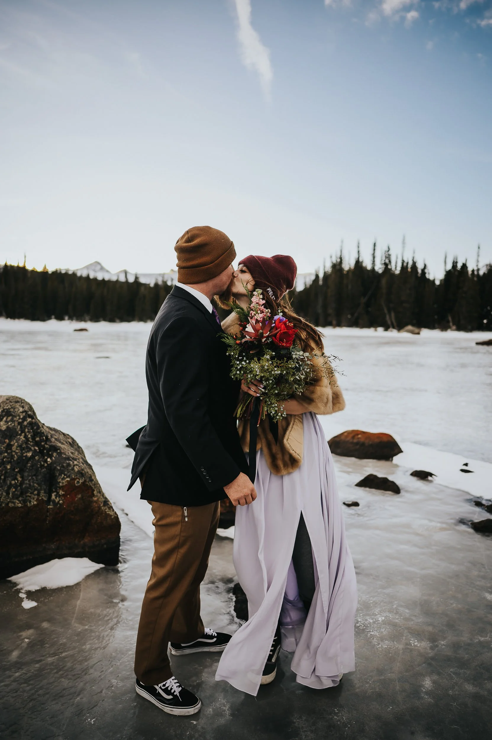 A groom leans toward his bride near a rushing Colorado mountain stream, her bouquet in hand.