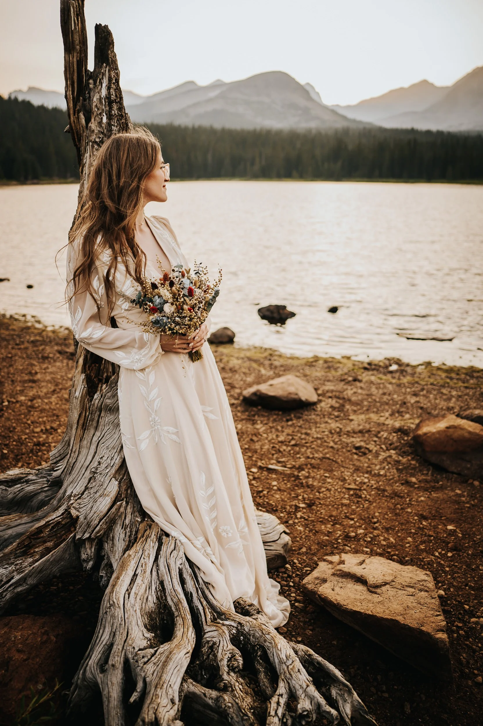 A bride in a boho lace dress stands at the edge of a Colorado lake, bouquet in hand, fall foliage around her.