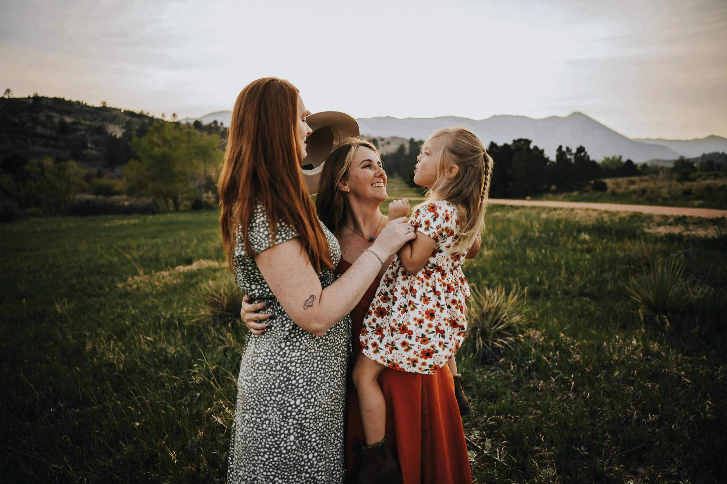 LGBTQ mothers hold their daughter between them in a grassy field at Ute Valley Park as the sun sets.