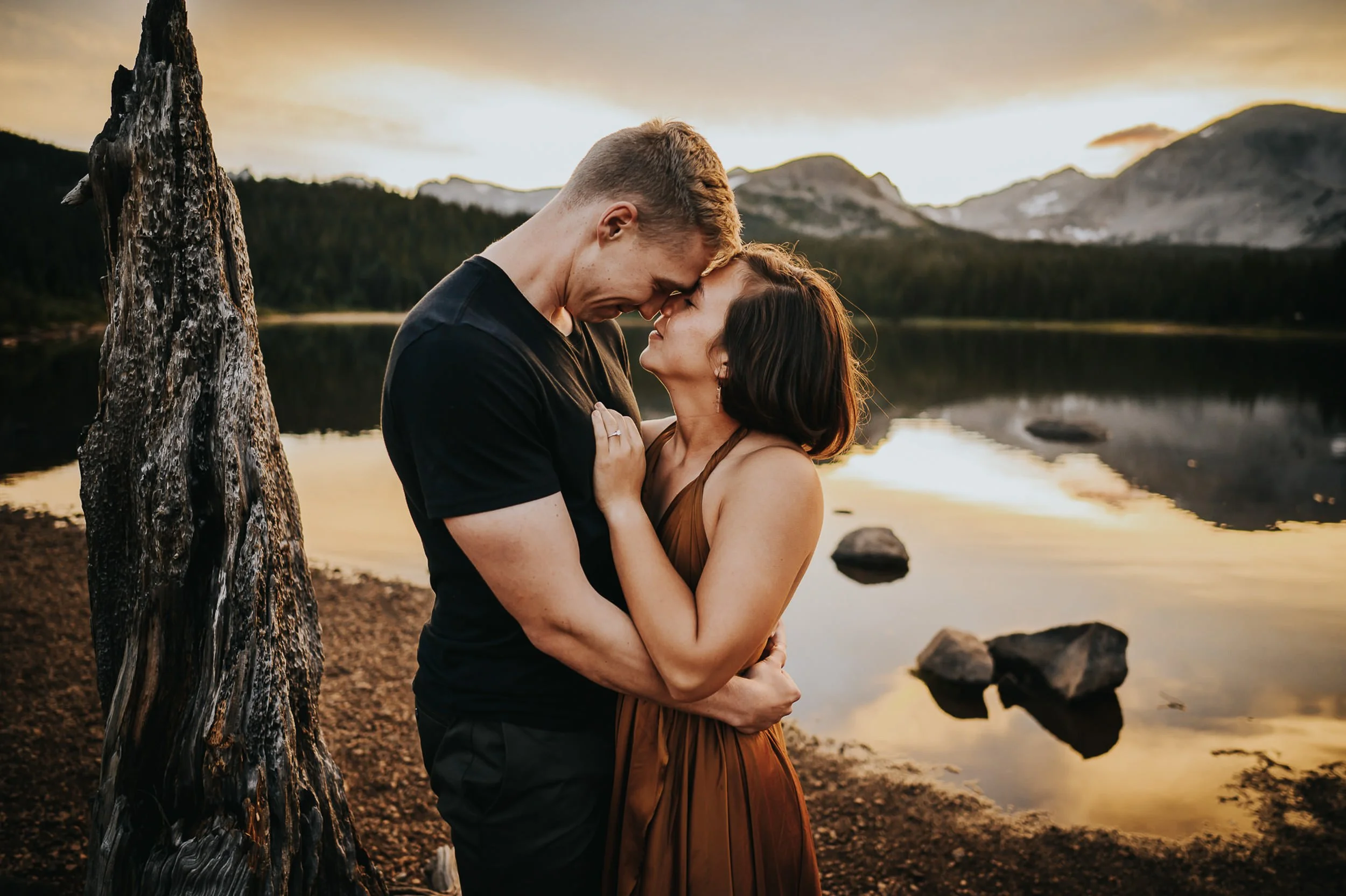 A couple leans into each other by a Colorado alpine lake, he in a black shirt, she in a rust dress, foreheads nearly touching.
